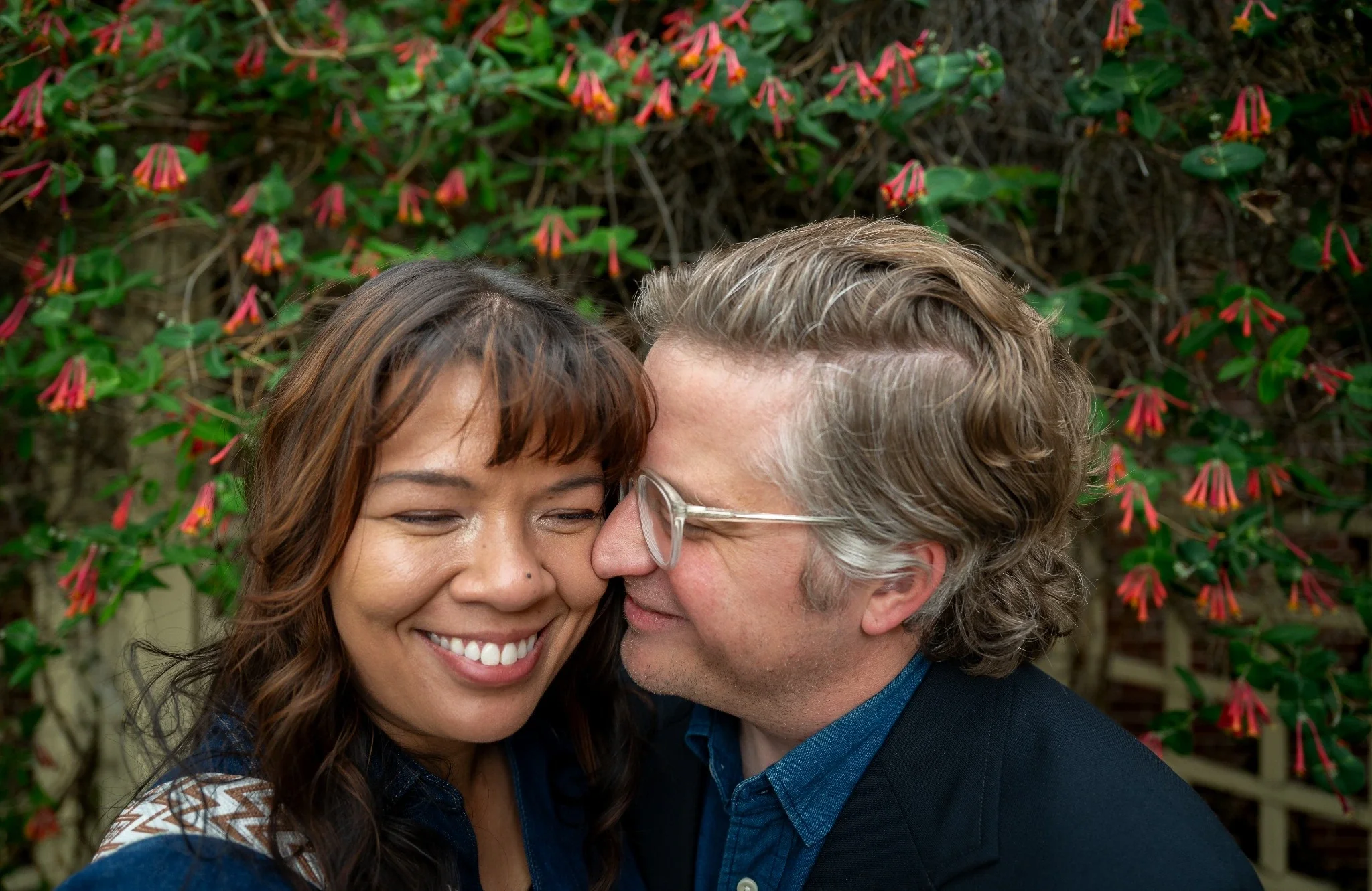 Intimate couples portrait in Olde Town Arvada, Colorado of a smiling couple embracing beneath flowering vines during a relaxed downtown engagement session by Ryan Kost Photography.