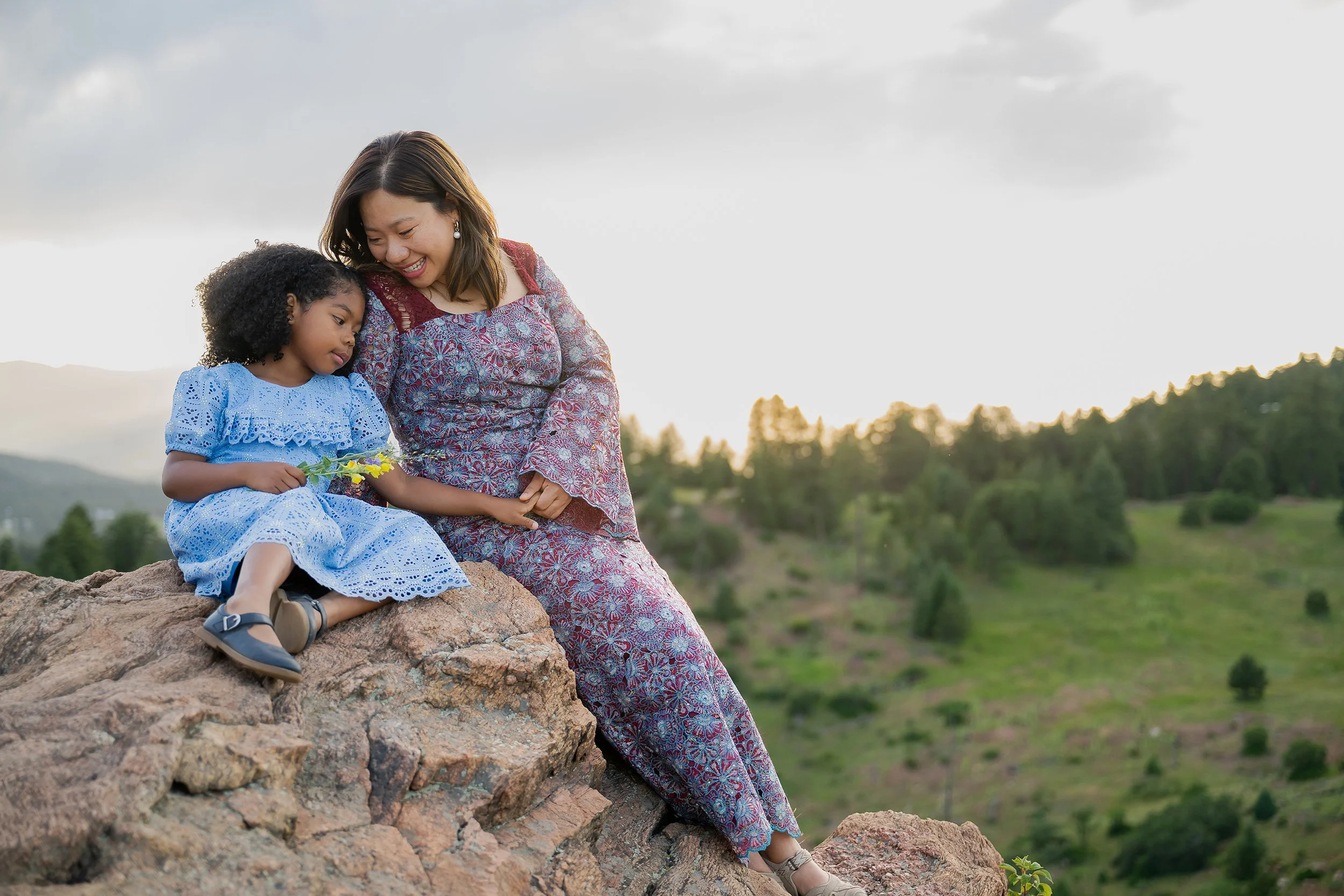 A soft, tender and loving moment between mom and daughter sitting on rocks at Mount Falcon West Trailhead in Colorado during a family photoshoot captured by Ryan Kost