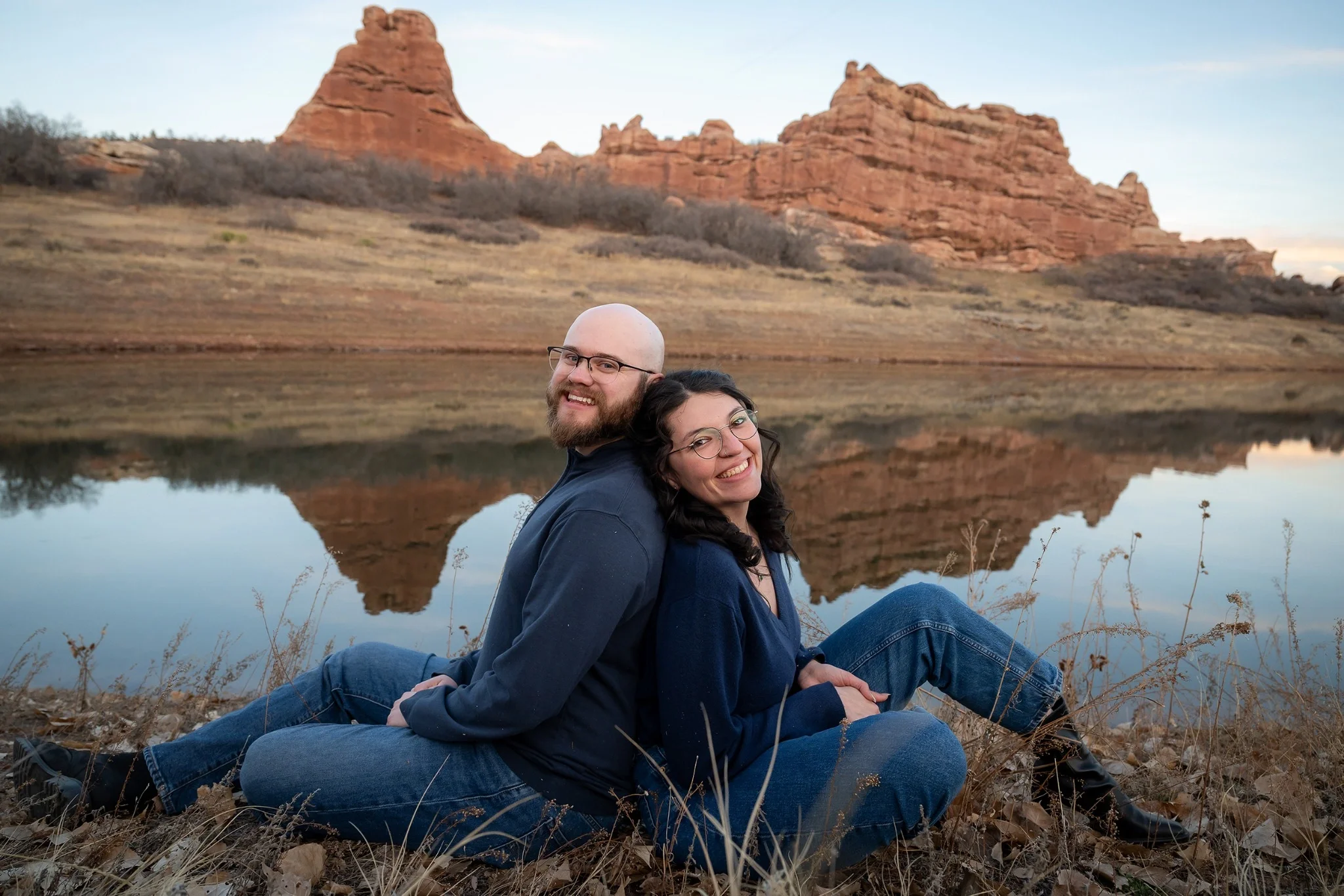 Engagement portraits of a couple sitting back to back by a reflective pond at South Valley Park with red rock formations in Littleton, Colorado by Ryan Kost