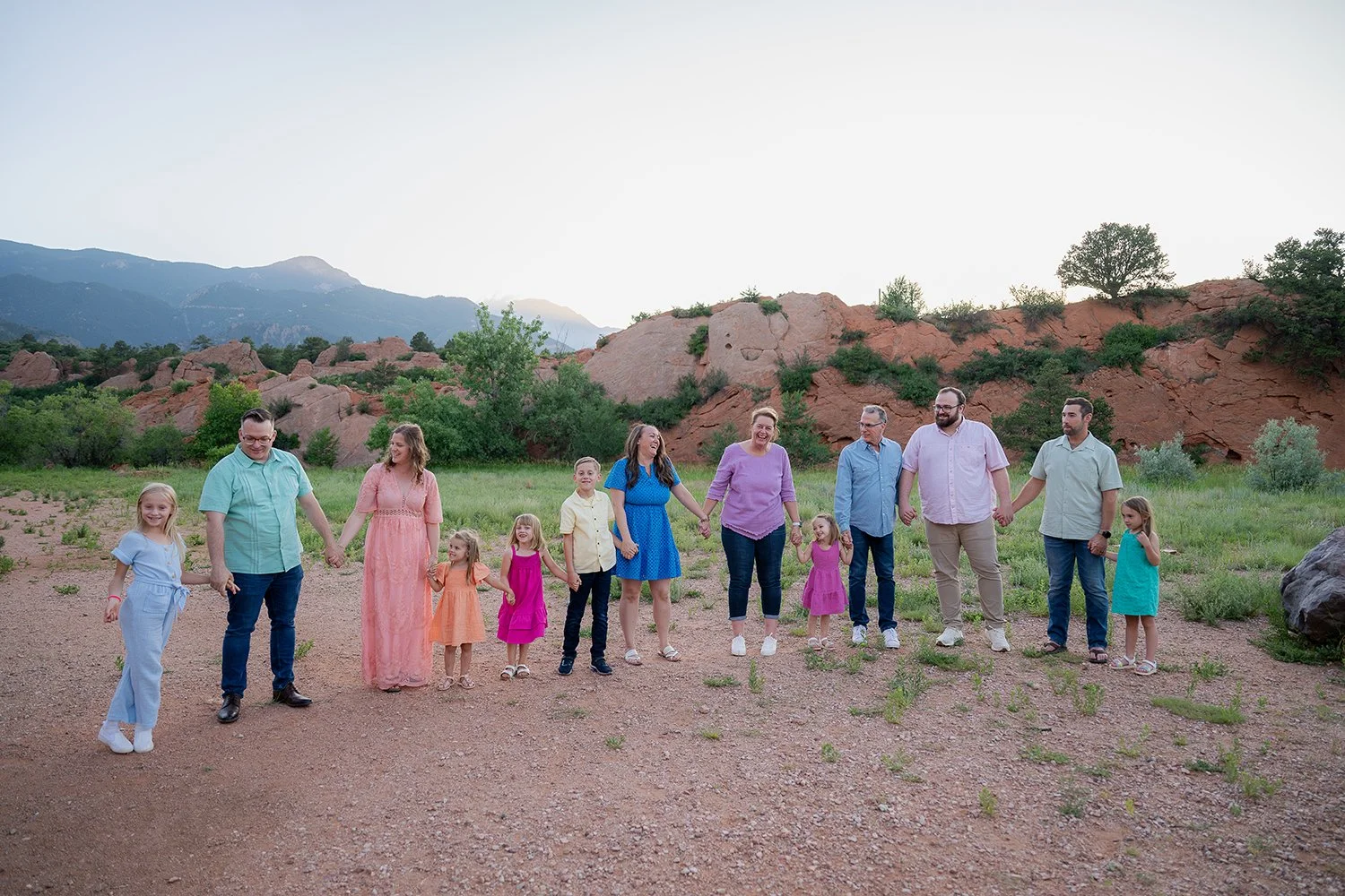 Multigenerational family holding hands walking together at Red Rock Canyon Open Space with red rock cliffs and Pikes Peak in the background.