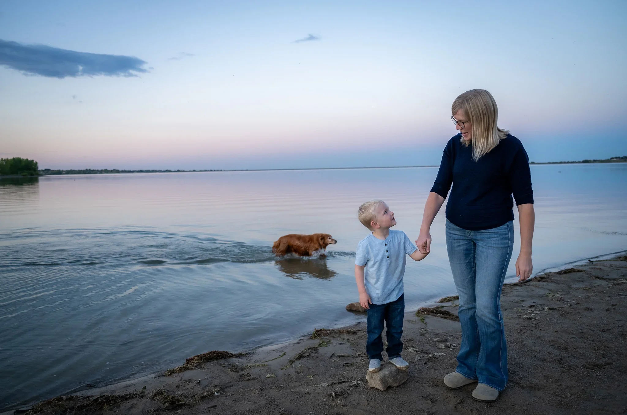 Mom and son hold hands while dog swims in water at Standley Lake in Westminster, Colorad.  Relaxed, natural family photography captured by Ryan Kost.