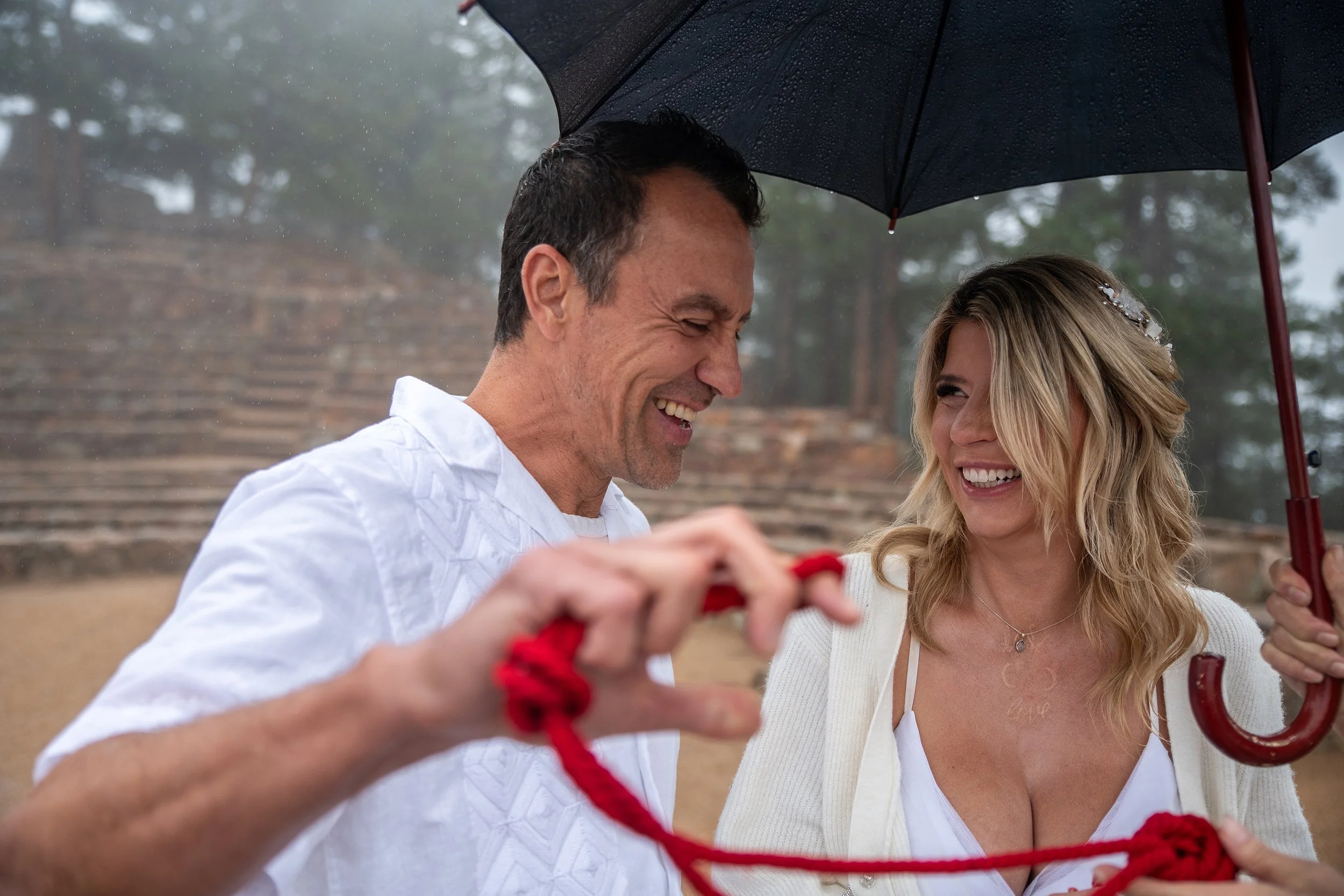 A man and woman smiling under an umbrella during a rainy day. The man is holding a red rope, and they are outdoors with trees and stairs in the background.