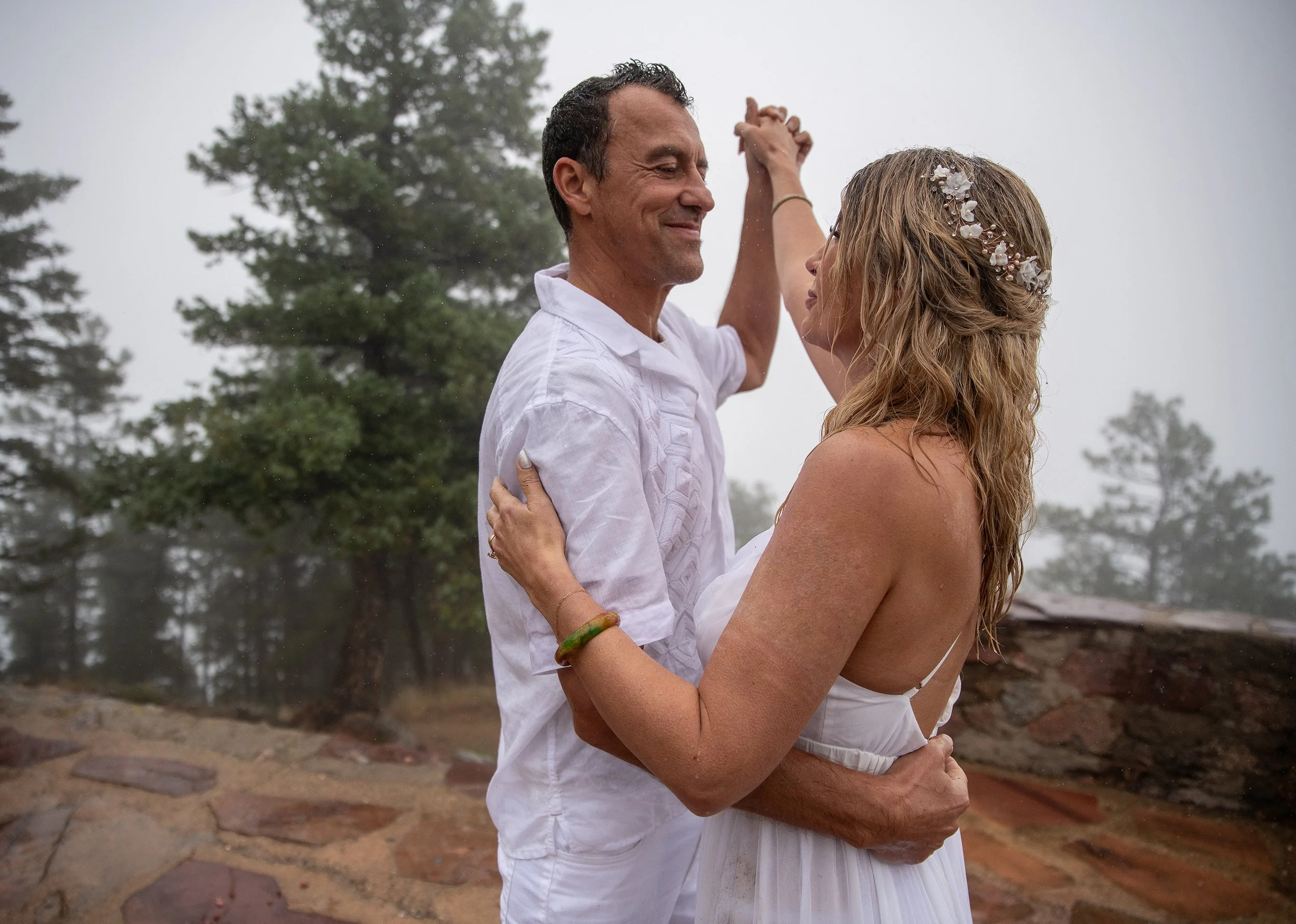 A couple dancing outdoors on a foggy day, with trees in the background. The man is smiling, wearing a white shirt, and the woman is wearing a white dress with a floral headpiece.