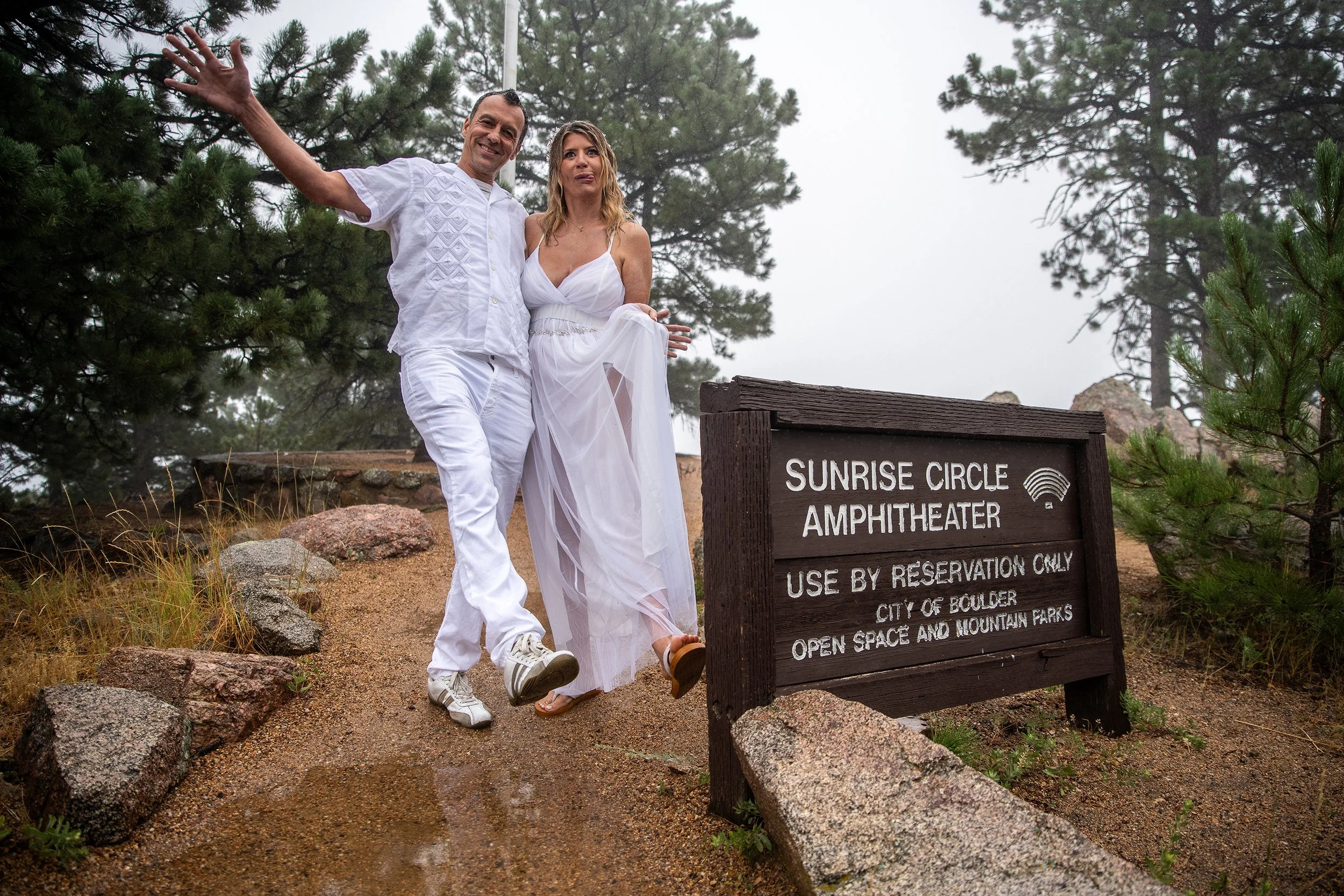 A man and a woman dressed in white standing near a sign that reads 'Sunrise Circle Amphitheater, Use by reservation only, City of Boulder, Open space and mountain parks' in a forested area.