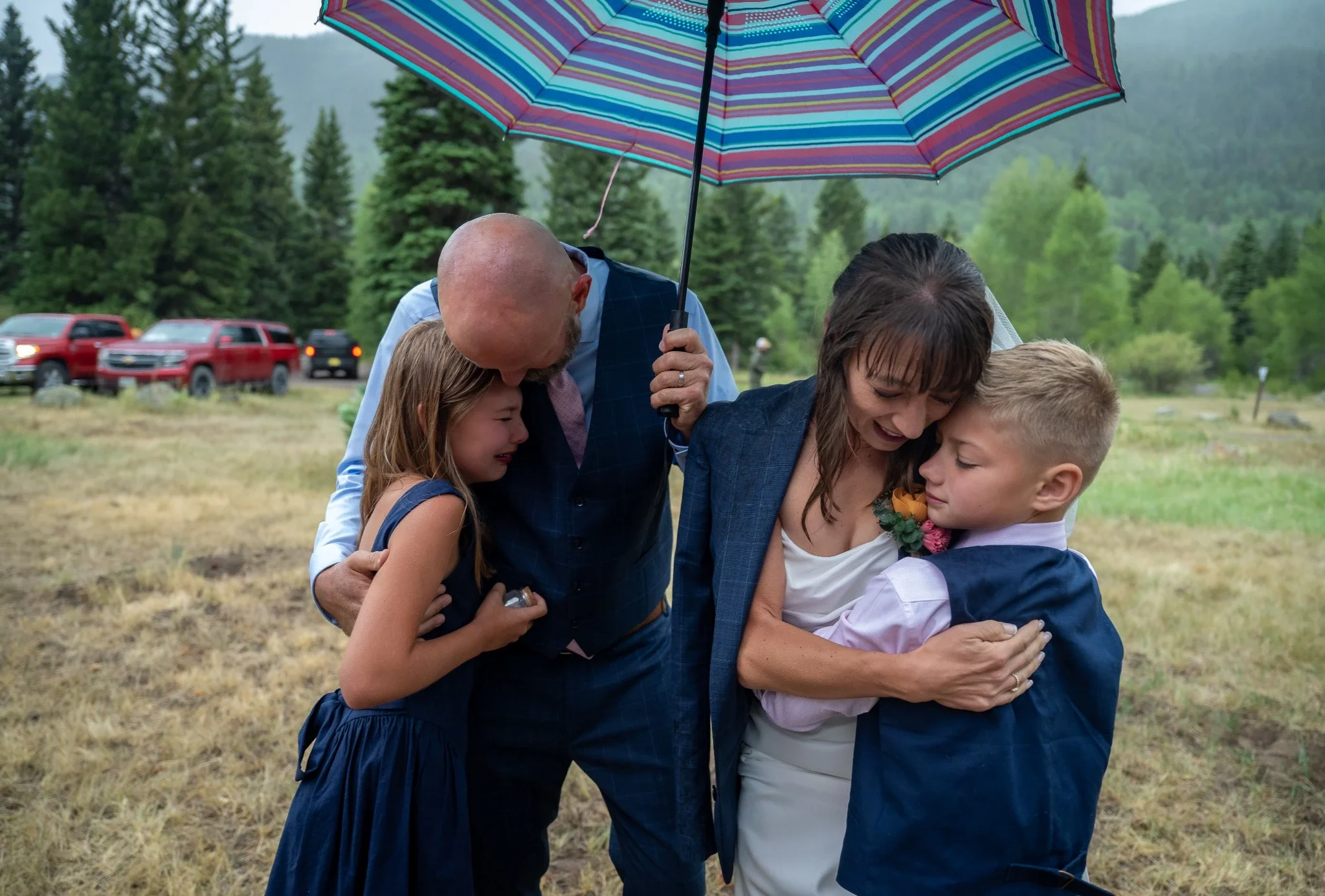 Emotional family embrace beneath a colorful umbrella during a rainy outdoor wedding ceremony in Horca, Colorado.