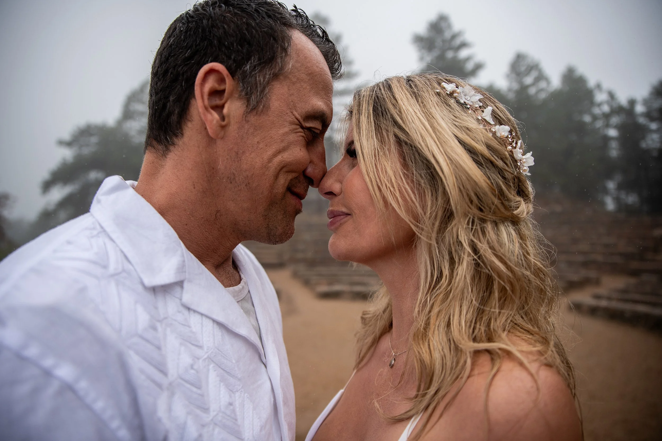 A man and woman standing face to face outdoors, touching foreheads and smiling softly. The woman has long blonde hair with a floral headband and is wearing a white dress. The man has dark hair and is wearing a white shirt. The background features blu