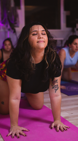 A woman practicing yoga in a studio, in a cow pose with back arched, with other women in the background.