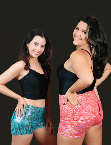 Two women smiling, standing side by side against a dark background, wearing black tops and colorful shorts.