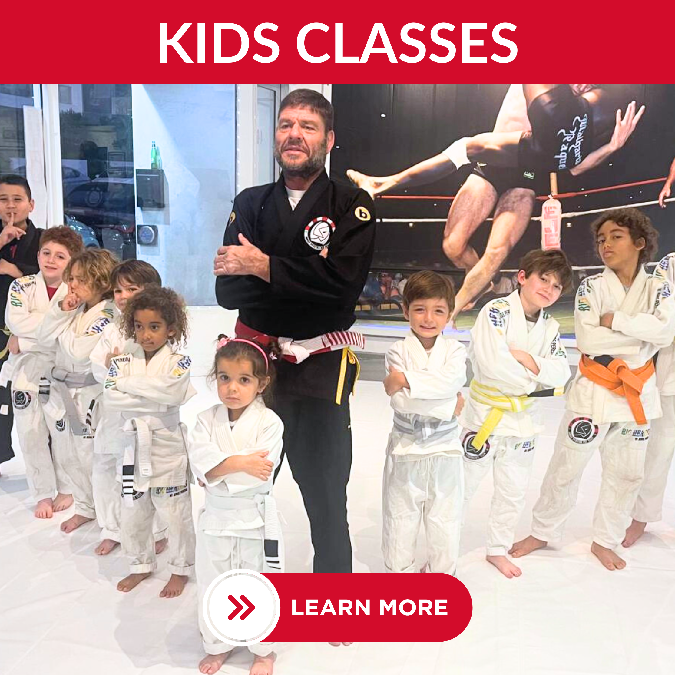 A group of children in white judo uniforms with colored belts posing with an instructor in a martial arts studio. The studio has a large poster of a mixed martial arts fight in the background and a red banner with white text that says 'Kids Classes'.
