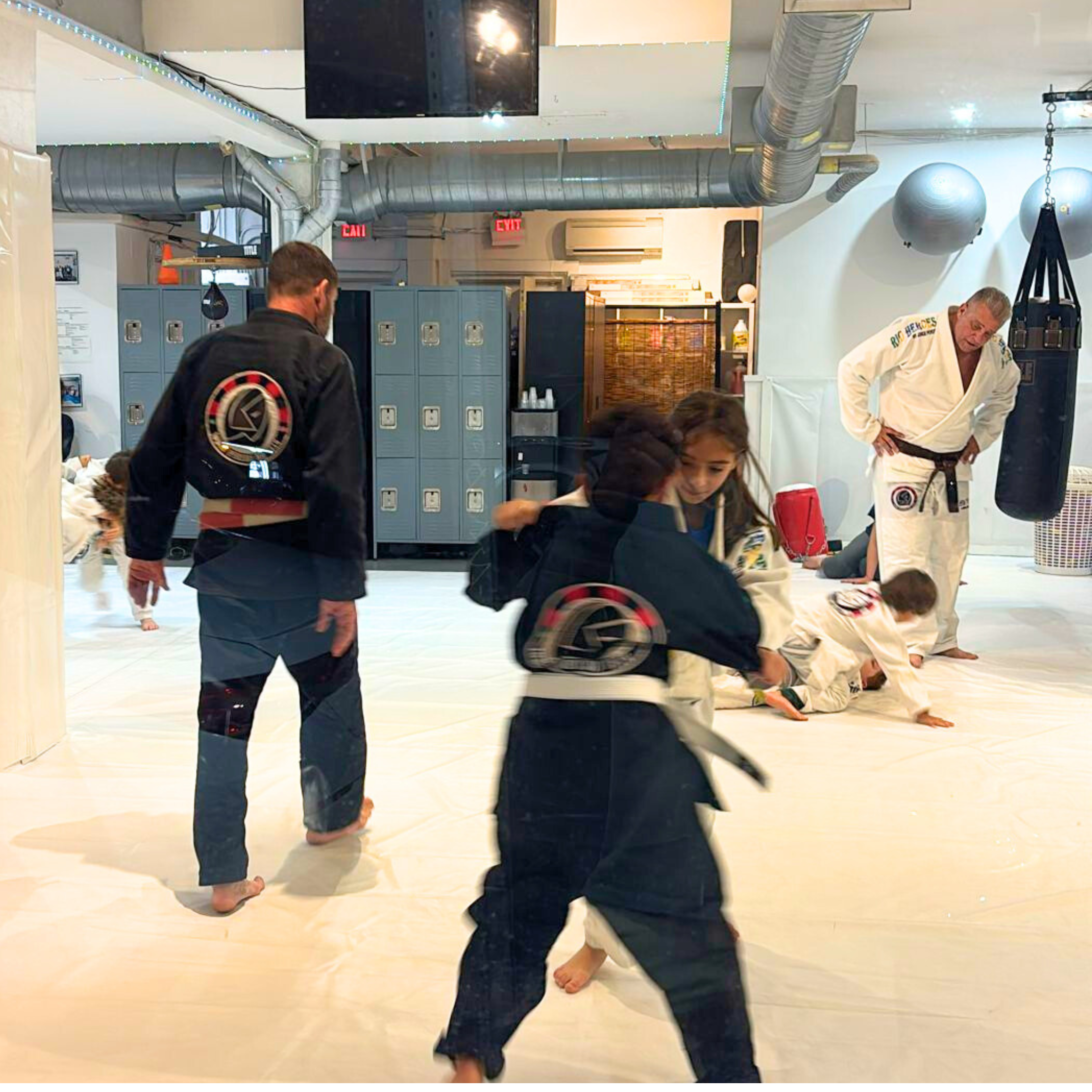 Children practicing Brazilian Jiu-Jitsu in a gym, wearing martial arts uniforms with a logo on the back, with an instructor in a white gi and black belt in the background.