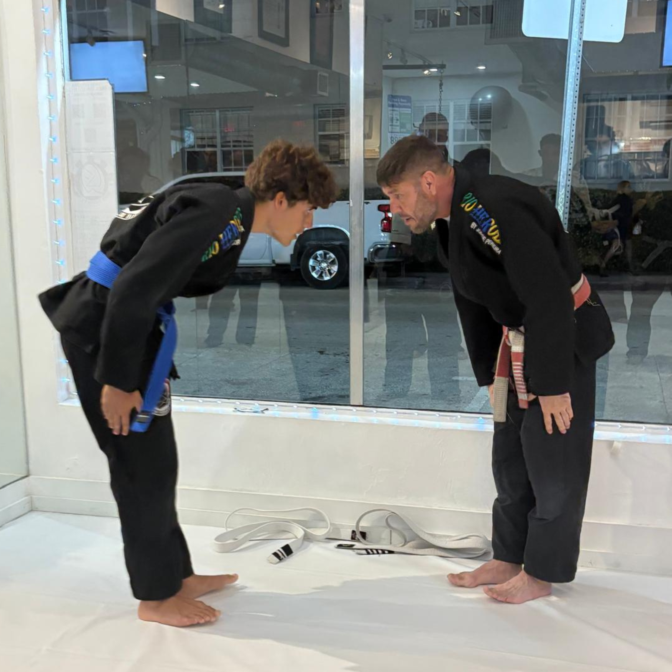Two people in Brazilian Jiu-Jitsu gis bowing to each other inside a gym, with a white mat and an orange and white belt lying on the mat.