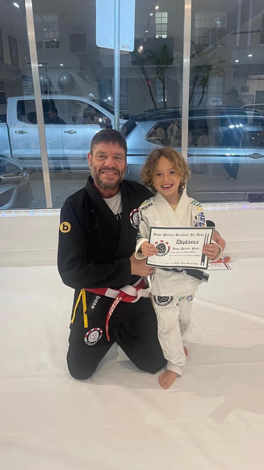 A man and a young boy in Brazilian Jiu Jitsu uniforms posing inside a martial arts gym, with the boy holding a diploma, both smiling.