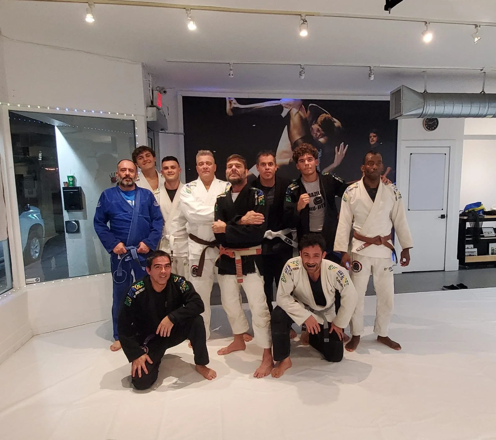 Group of people in Brazilian Jiu-Jitsu gis posing in a martial arts gym. Some are standing while others are kneeling or crouching, with a large martial arts poster on the wall behind them.