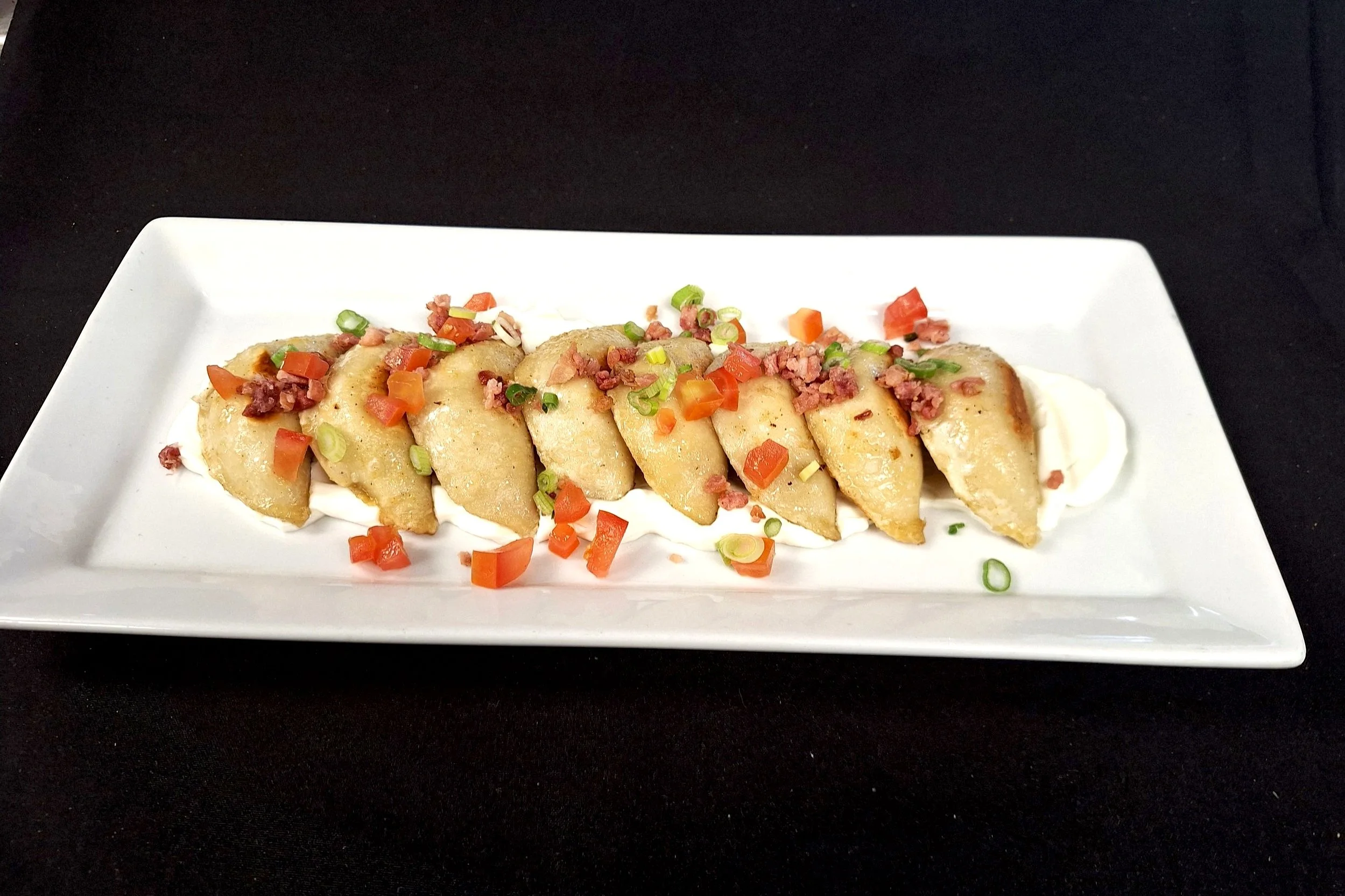 Close-up of five pieces of beef on a crispy wonton chip, topped with green onions, drizzled with brown sauce, served on a white rectangular plate with a drizzle of creamy sauce underneath.