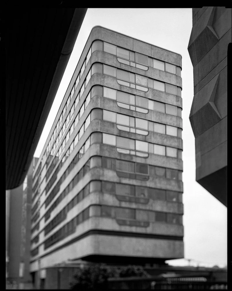 Un immeuble moderne en béton et verre, vu en contre-plongée en noir et blanc, avec des fenêtres arrondies et des balcons, situé entre deux autres bâtiments. Architecture Bordeaux