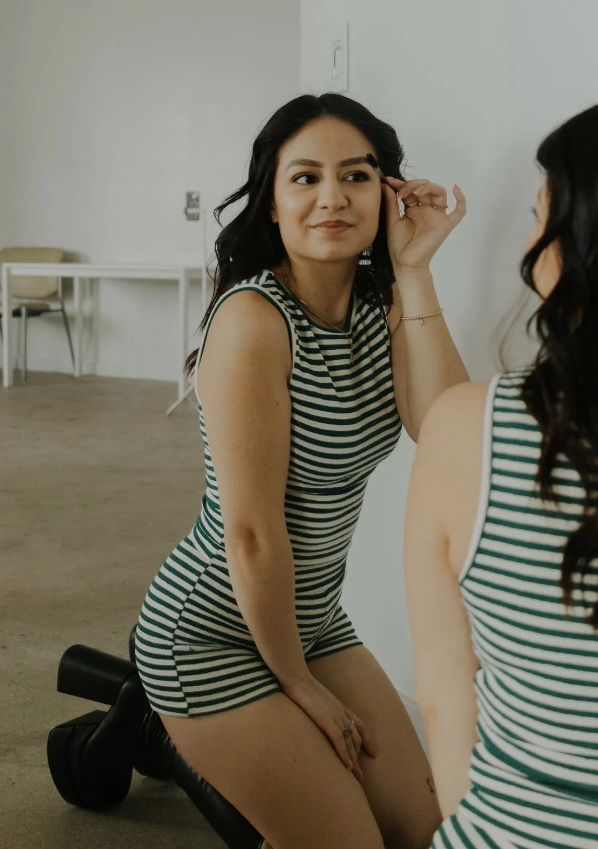 A young woman with long dark hair taking a selfie indoors. She is wearing a dark top and has a subtle smile. The background includes a window with blinds.