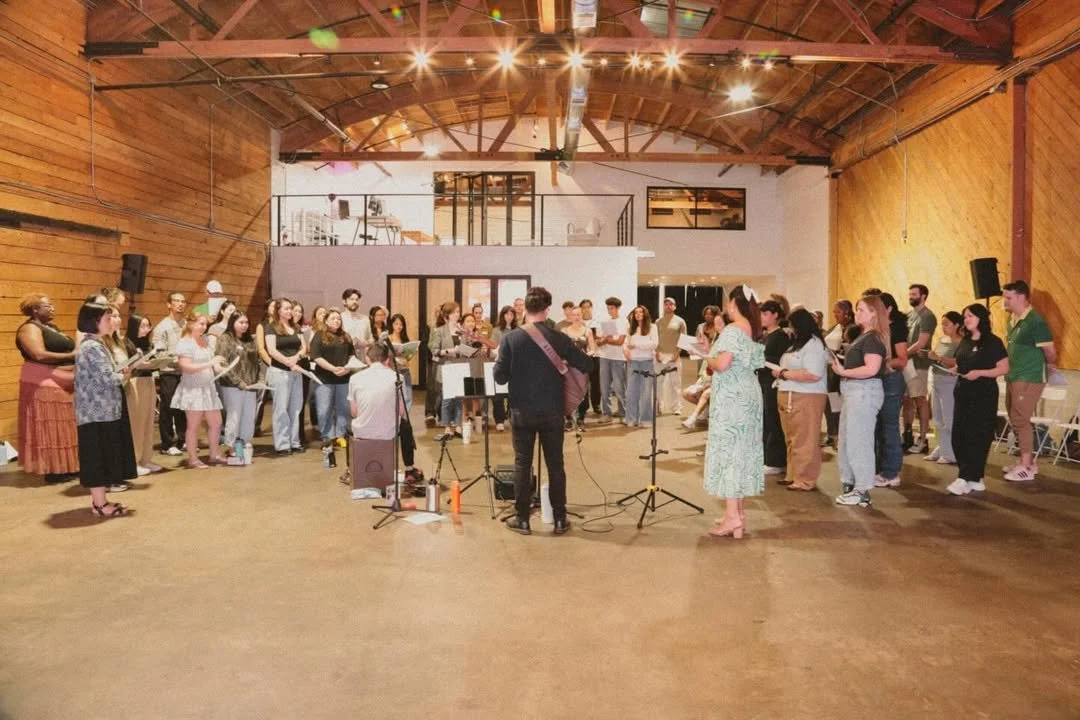 A group of people gathered in a spacious, wooden-finished hall, singing or rehearsing with a conductor and musicians, illuminated by overhead lights.