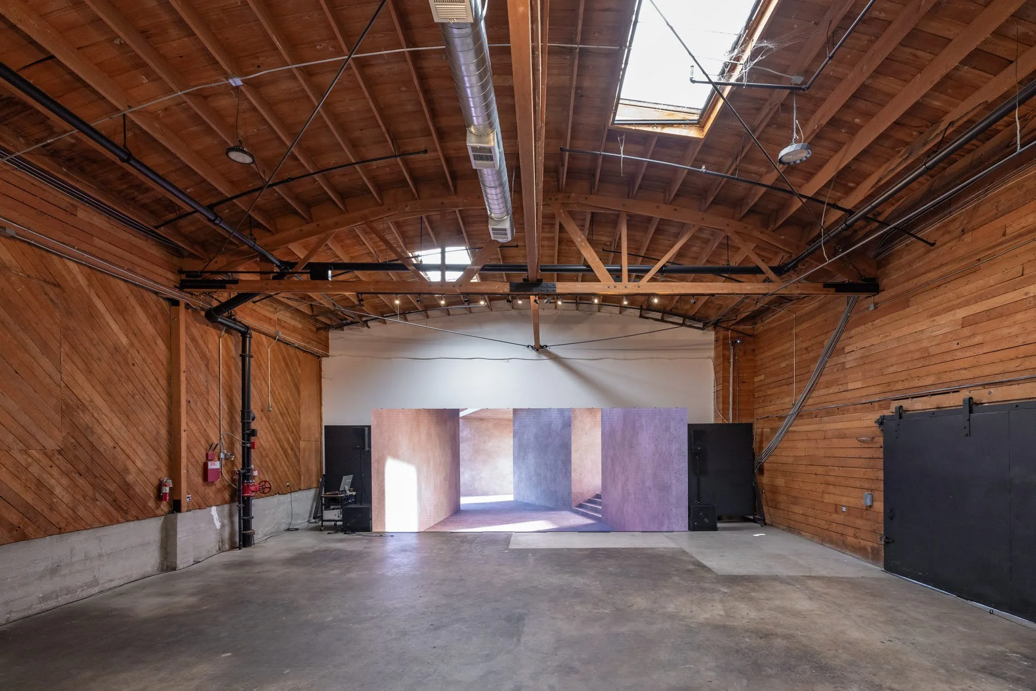 An empty interior space with wooden walls and ceiling, natural light coming through a skylight, and a stage at the back.
