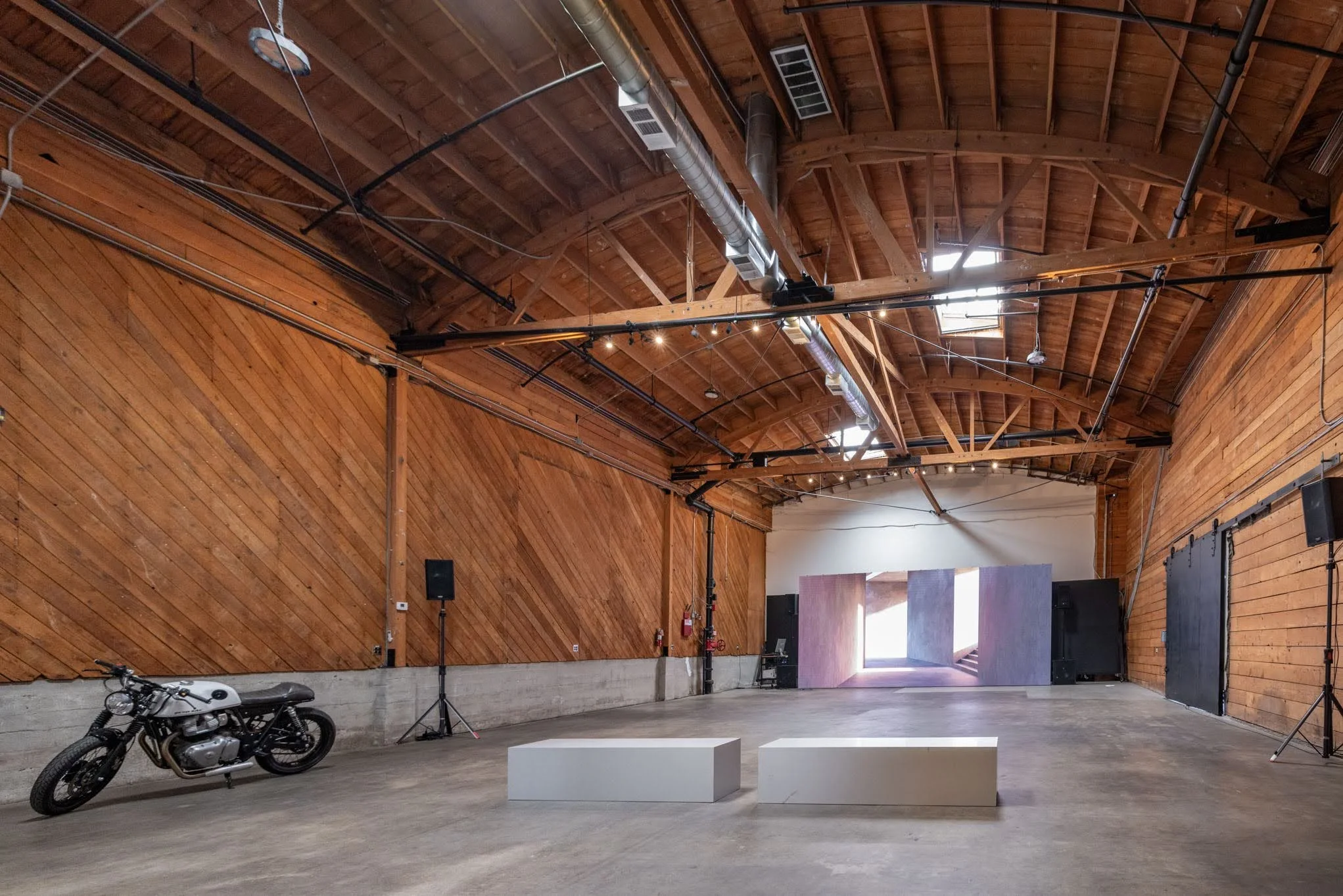 An indoor space with wooden walls and ceiling, a motorcycle parked on the left side, two white rectangular pedestals in the center, black speakers on both sides, and lighting fixtures hanging from the ceiling. There is an open doorway with natural li