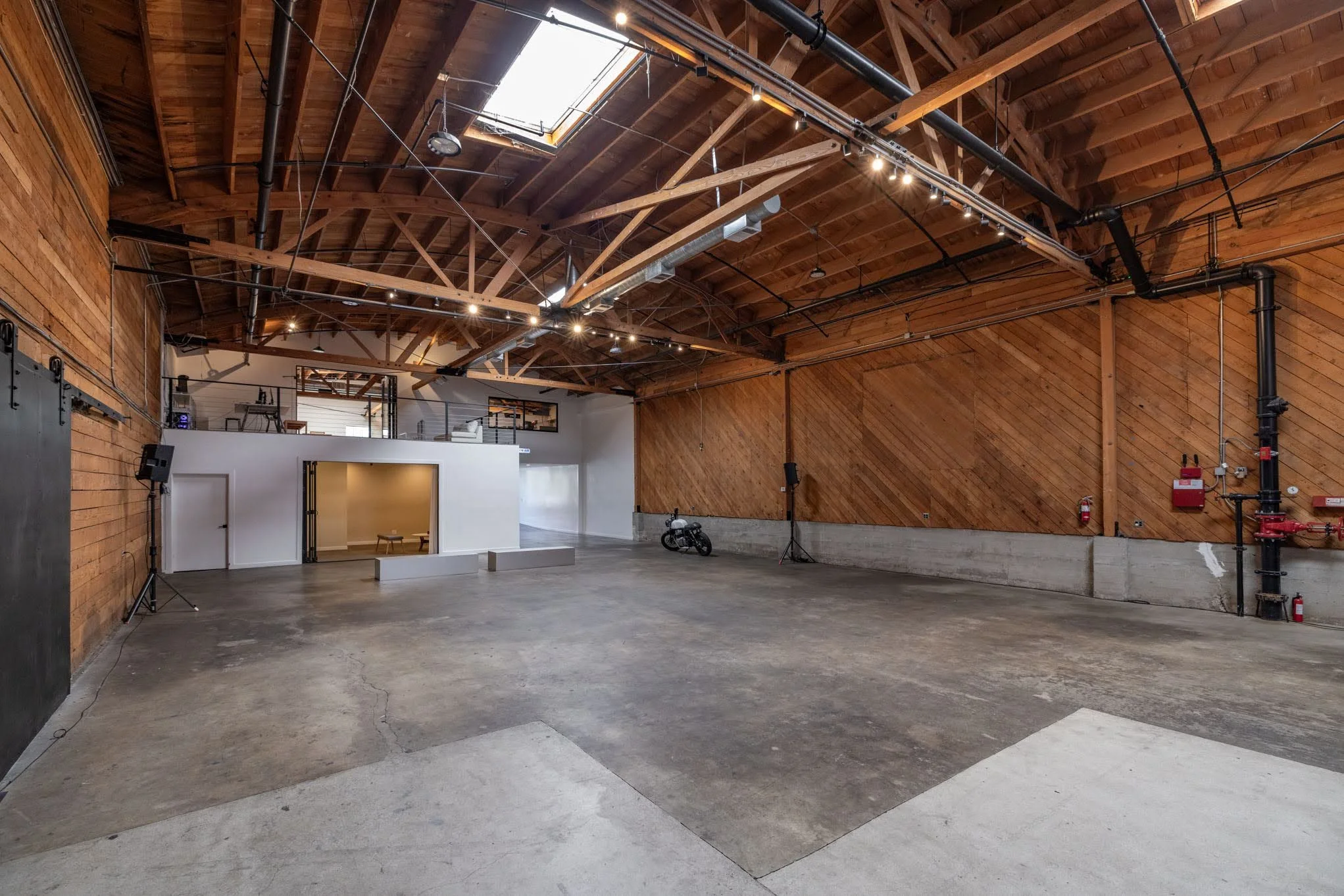 Large indoor space with wooden walls and ceiling, skylight, some lighting, and various equipment, including a bicycle, speaker, and fire safety gear.