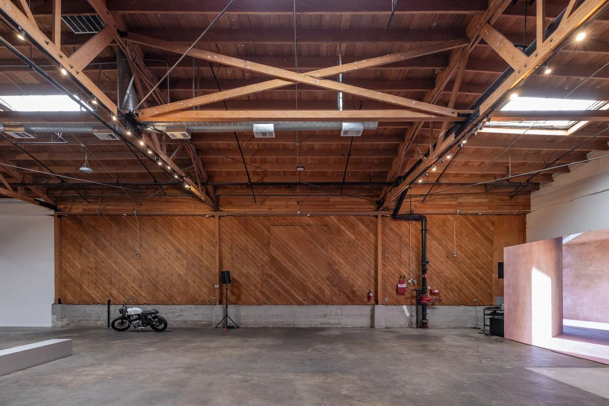 Interior of a large room with high wooden ceiling and exposed beams, a motorcycle parked on the floor, and medical equipment like red fire hose and black speaker on the right side.