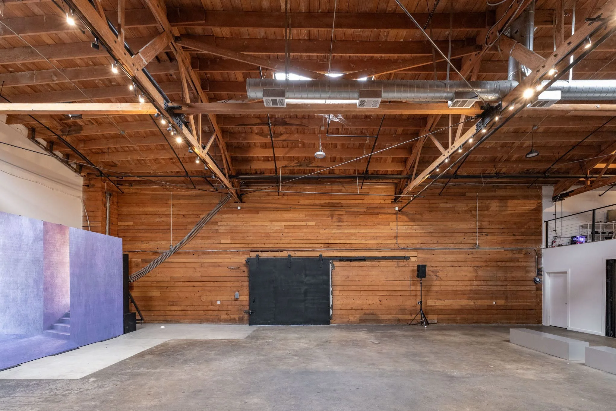 Empty indoor space with wooden ceiling beams, ductwork, and a large open floor area, likely a studio or event space.