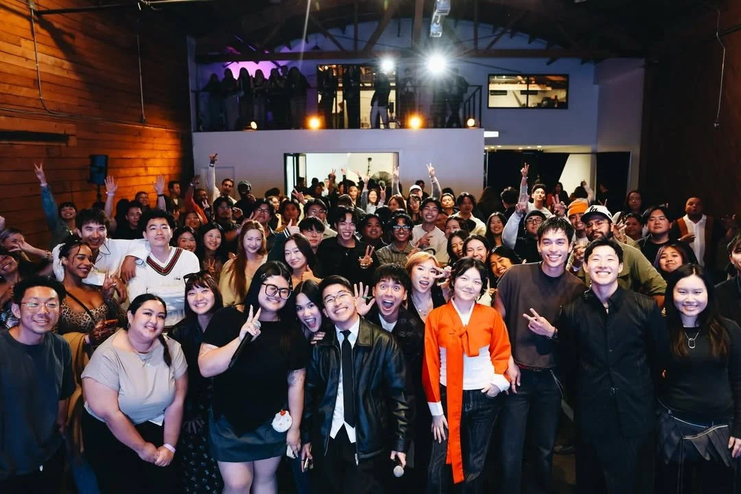 Large group of people gathered at an indoor event, smiling and posing for a photo, with some making peace signs and raising their hands.