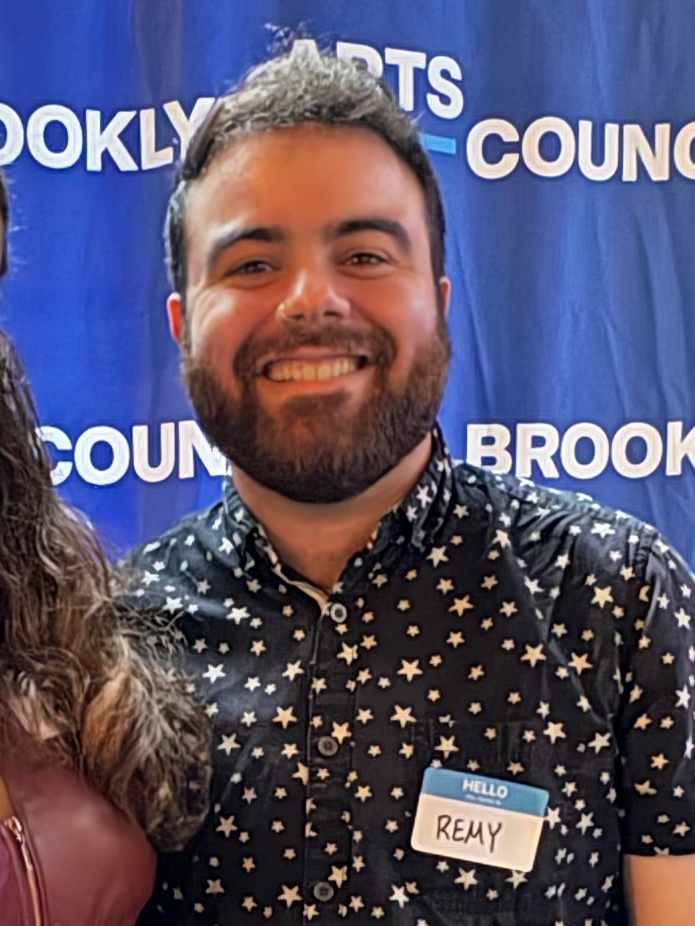 Smiling man with a beard, wearing a black shirt with white star patterns and a name tag that reads 'Remy', standing in front of a blue backdrop that says 'Brooklyn' and 'Council'.