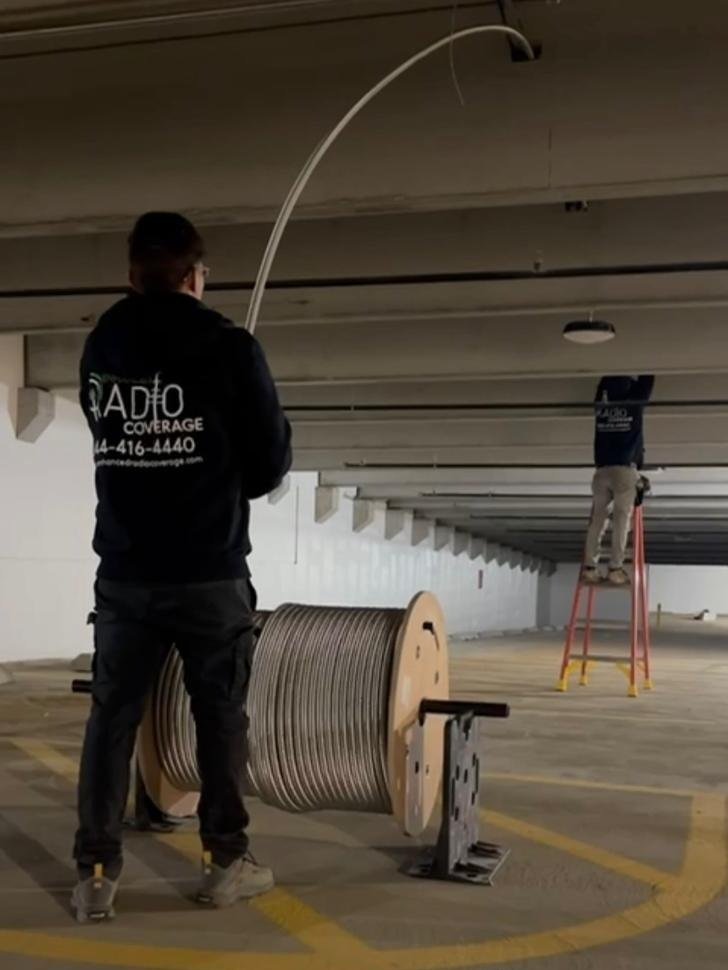 Two workers in black jackets with radio coverage logos are installing or repairing wiring in a parking garage. One worker is standing on the ground near a large spool of cable while the other is on a step ladder on the ceiling.
