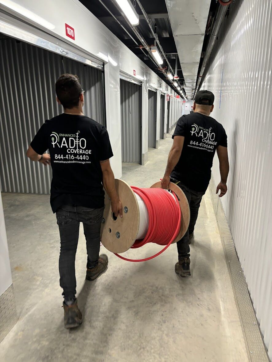 Two workers carrying a large spool of red cable through a corridor of storage units.