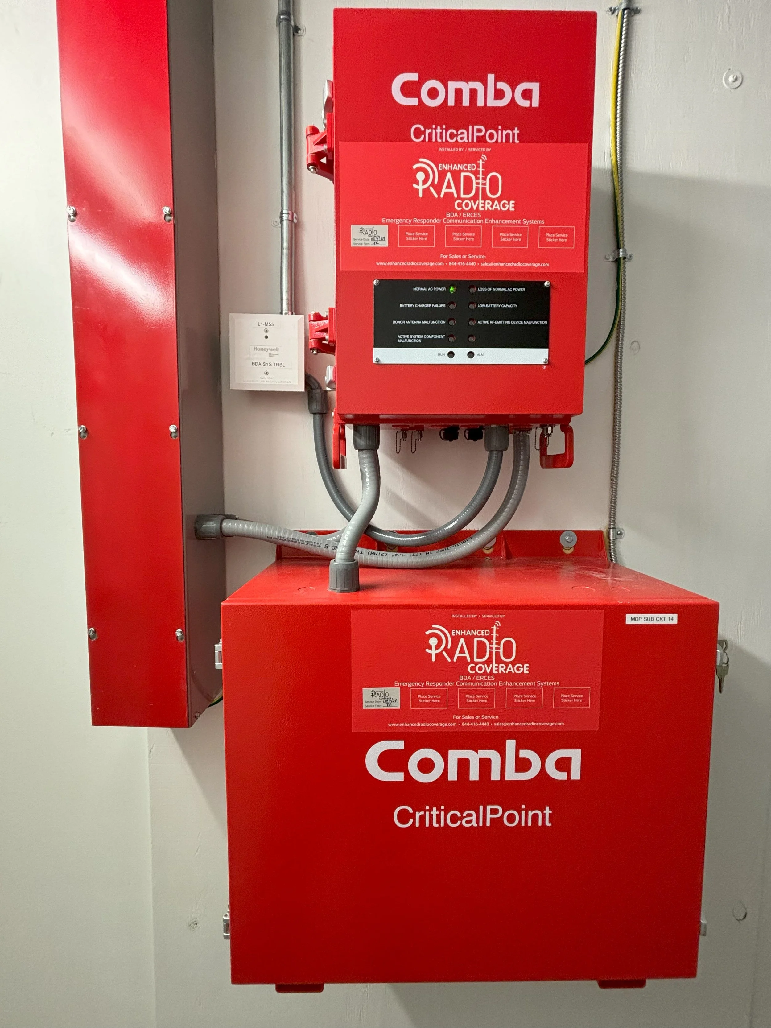 Red emergency radio communication system with labels, mounted on a wall, labeled 'Comba CriticalPoint' and 'Enhanced Radio Coverage'.