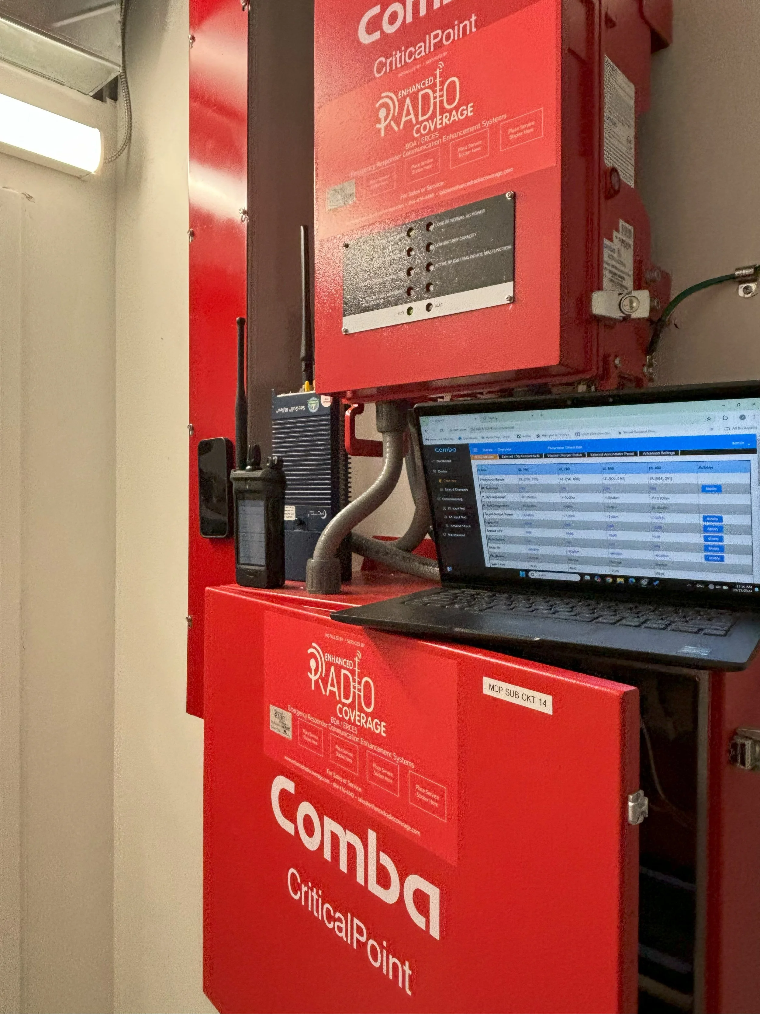 Red communication equipment with labels, a small handheld device, and a laptop displaying a spreadsheet, mounted on a wall.