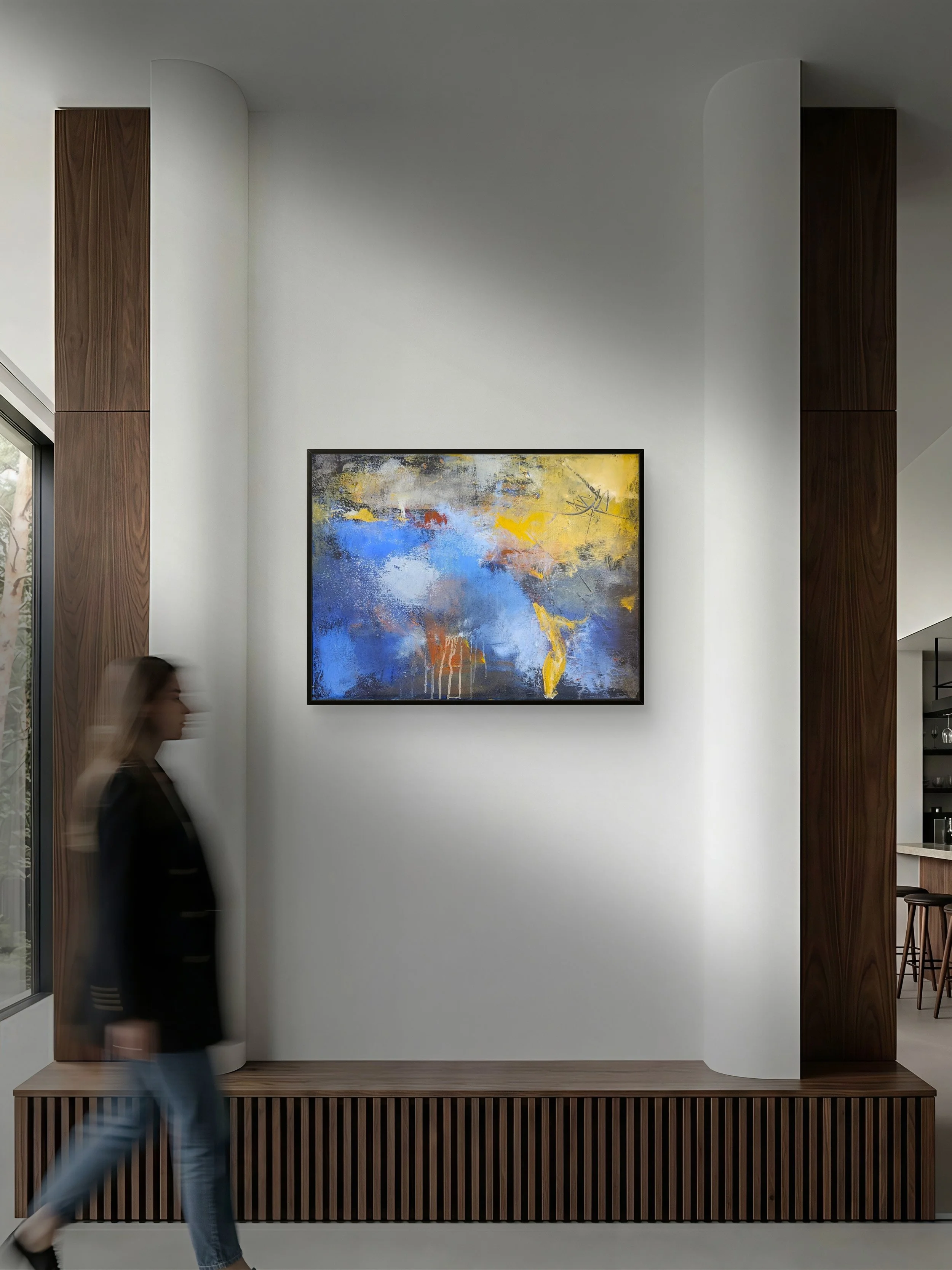 A modern interior with a white wall, a colorful abstract painting, wooden accents, and a blurred woman walking past.