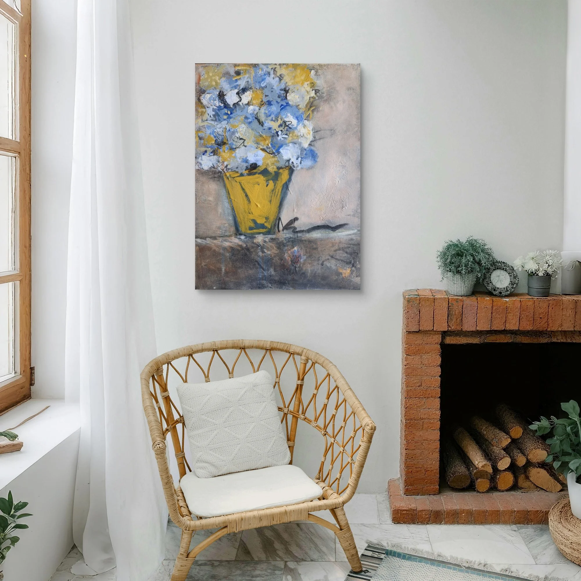 Wicker chair with a white pillow in front of a fireplace with logs, a brick hearth, houseplants, and a wall art of blue and white flowers in a yellow vase.