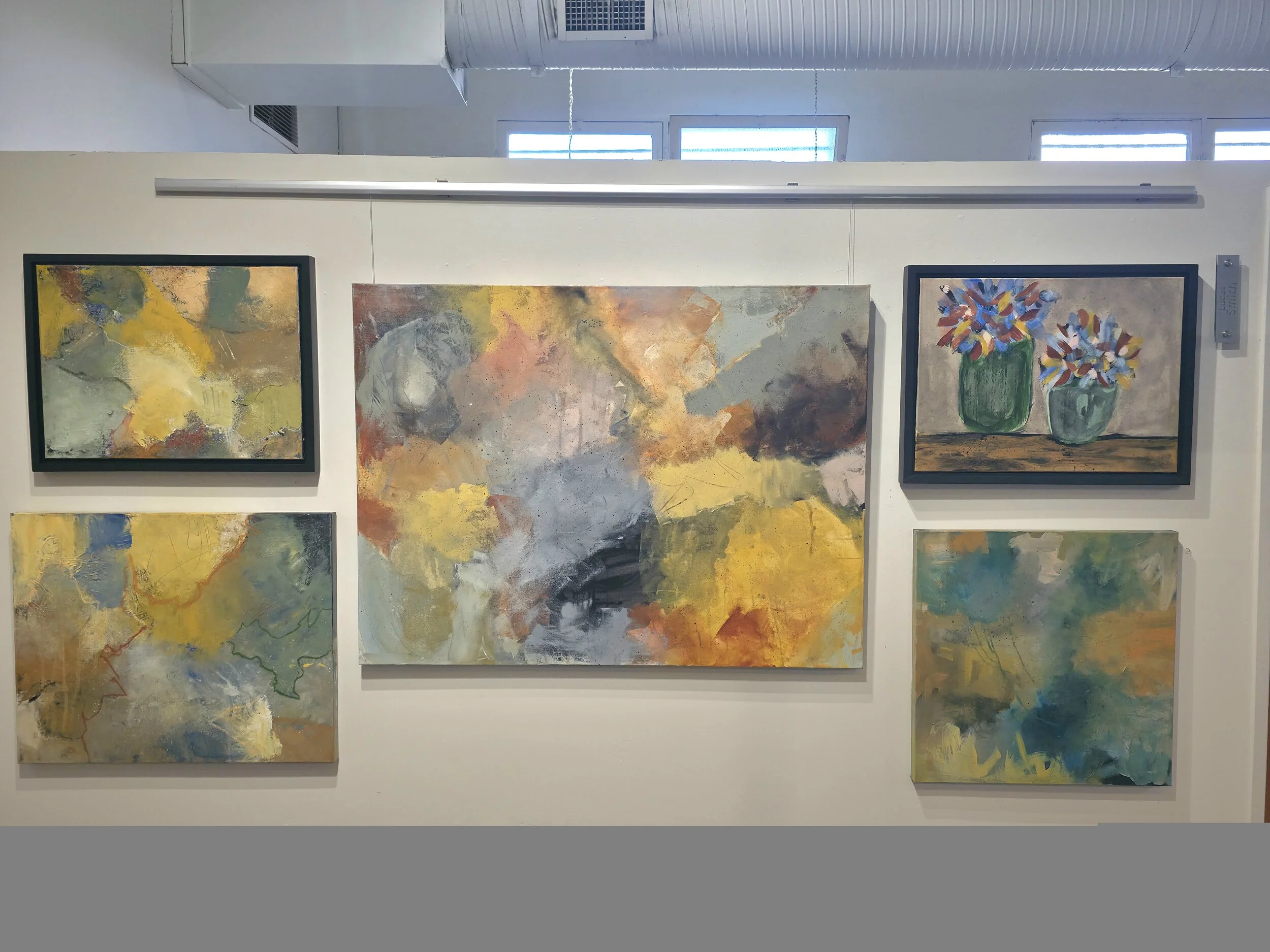 An art gallery display showcasing five abstract paintings on a white wall, with three of the paintings featuring colorful vases of flowers.