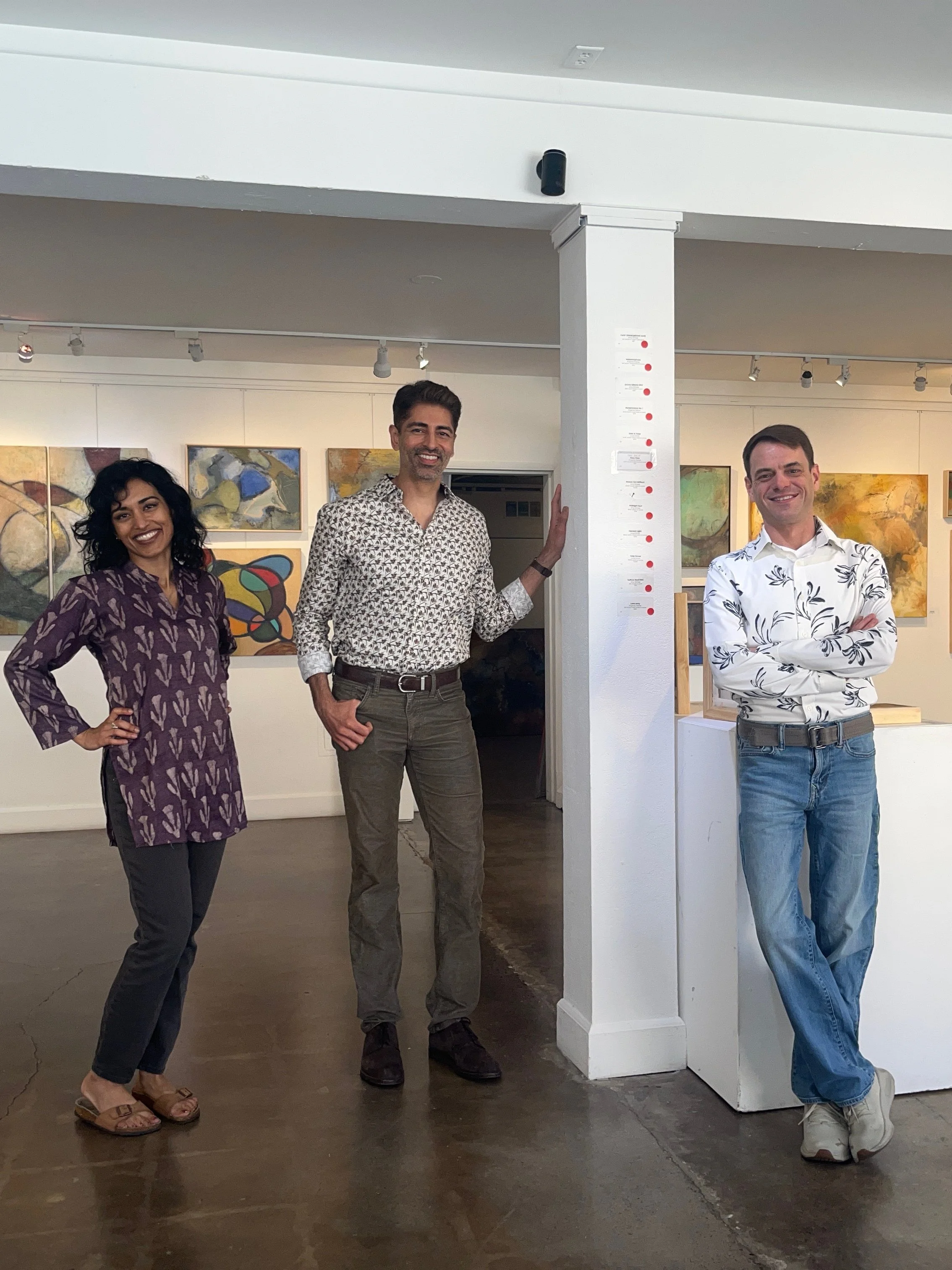 Three people standing inside an art gallery, next to a white wall with colorful paintings in the background. The woman on the left is wearing a purple patterned dress, the man in the middle is wearing a patterned shirt and brown pants, and the man on the right is wearing a white floral shirt and jeans. They are smiling and posing for the photo.