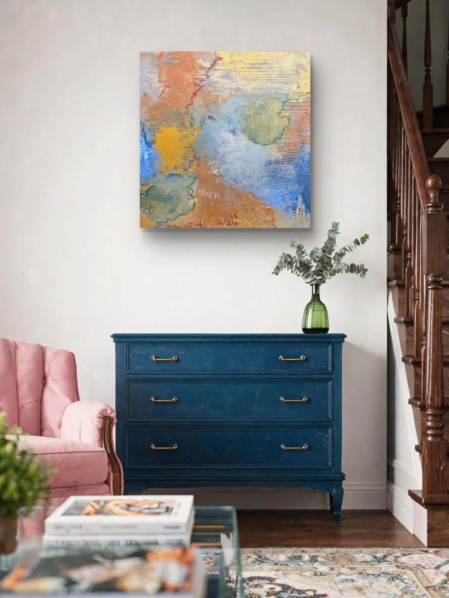 A blue dresser with gold handles in a living room, with a green vase with eucalyptus branches on top, pink sofa on the left, abstract colorful painting on the wall above, a patterned rug on the floor, and a wooden staircase on the right.