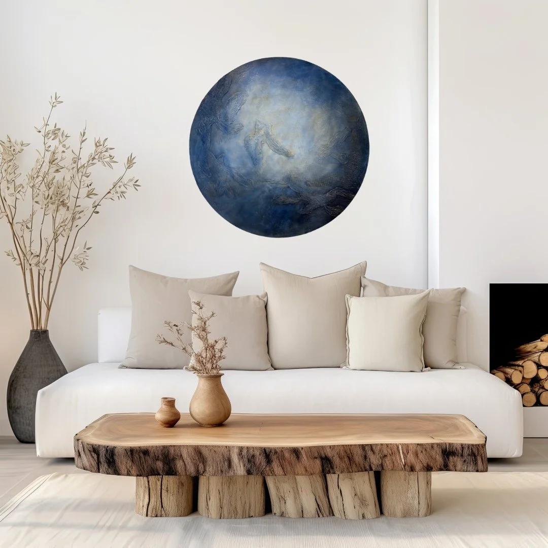 Minimalist living room with white sofa, beige cushions, a wooden coffee table, a large vase with branches, a small vase, abstract blue wall art, and a built-in fireplace with firewood.