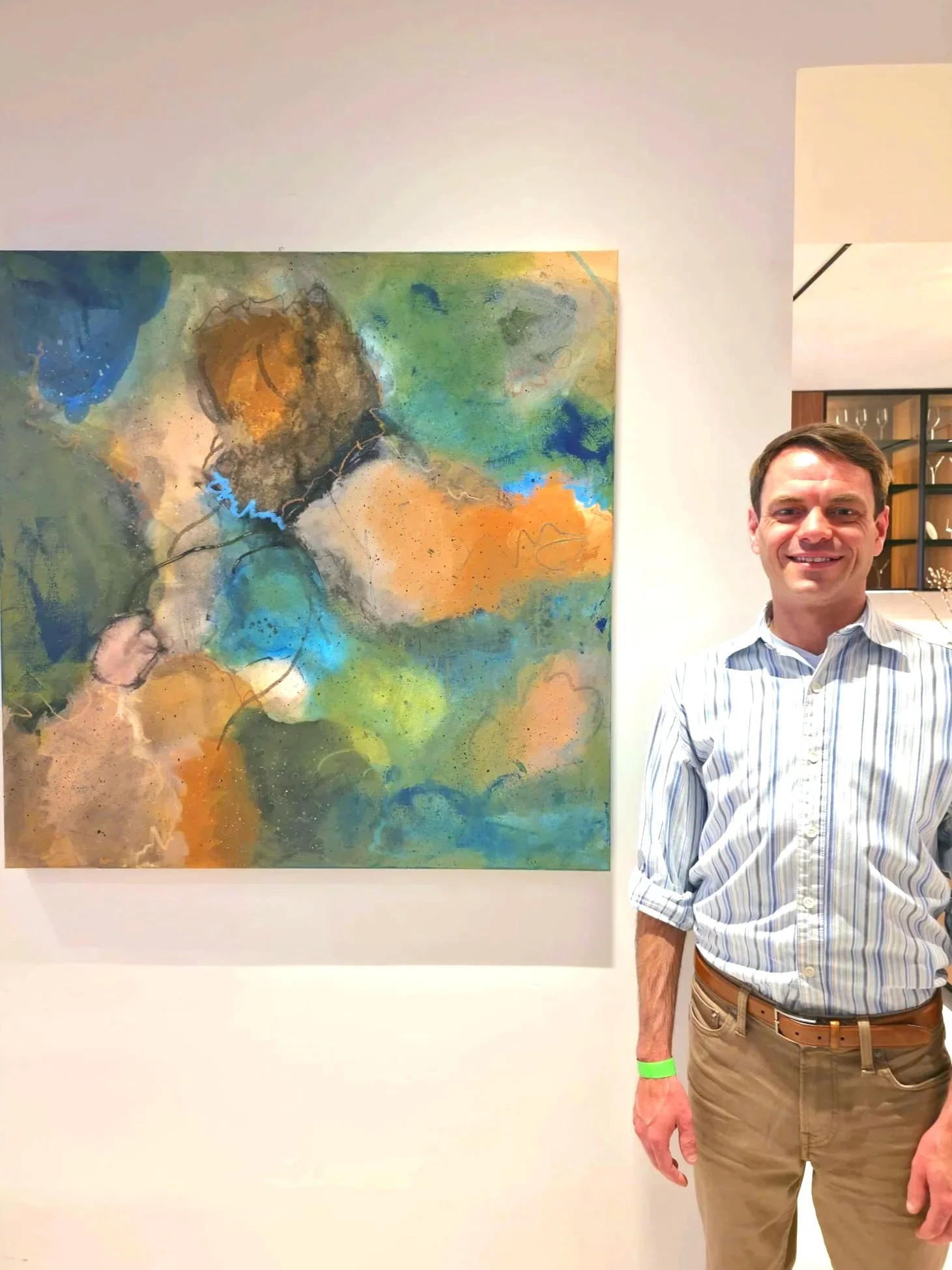 A man wearing a striped shirt and khaki pants stands in front of a colorful abstract painting at an art gallery.