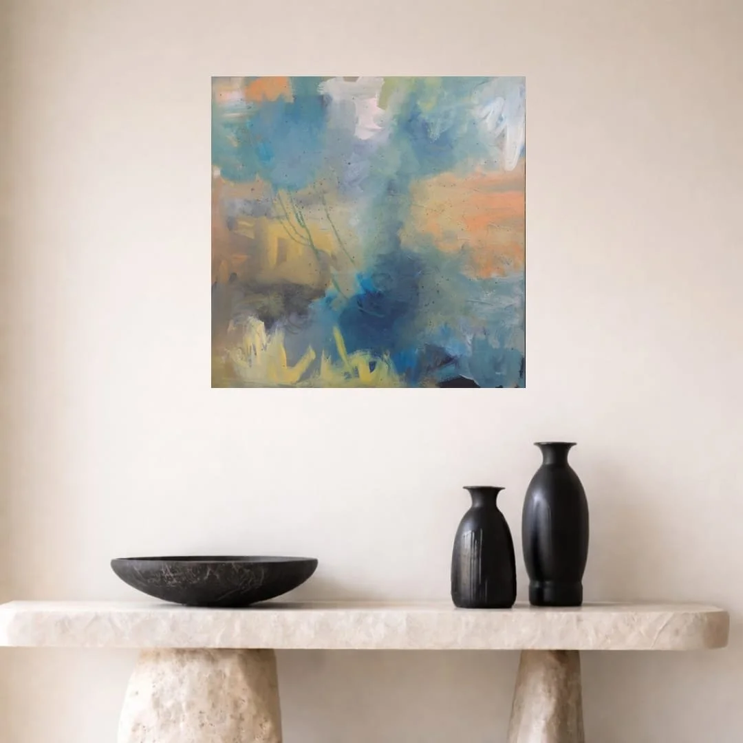 Abstract painting in shades of blue, yellow, gray, and peach hangs on a plain wall above a beige stone console table with black vases and a bowl.