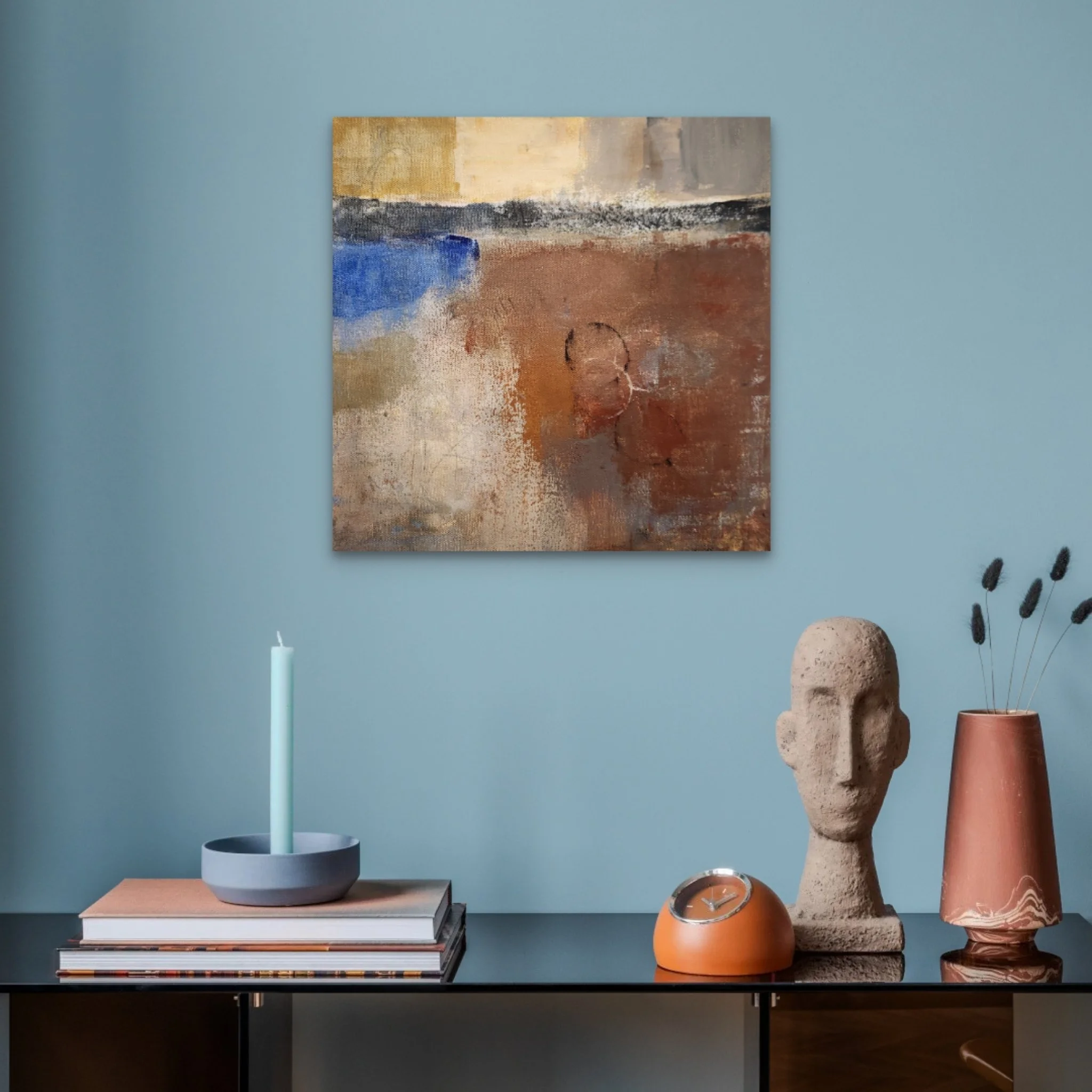 A display of modern decorative objects on a black metal table against a light blue wall, including a tall light blue candle in a black holder, stacked books, a gray bowl, a round orange clock, a large textured beige and brown sculpture, a pink vase, and a glass container with dried black plant stems, with an abstract painting on the wall.