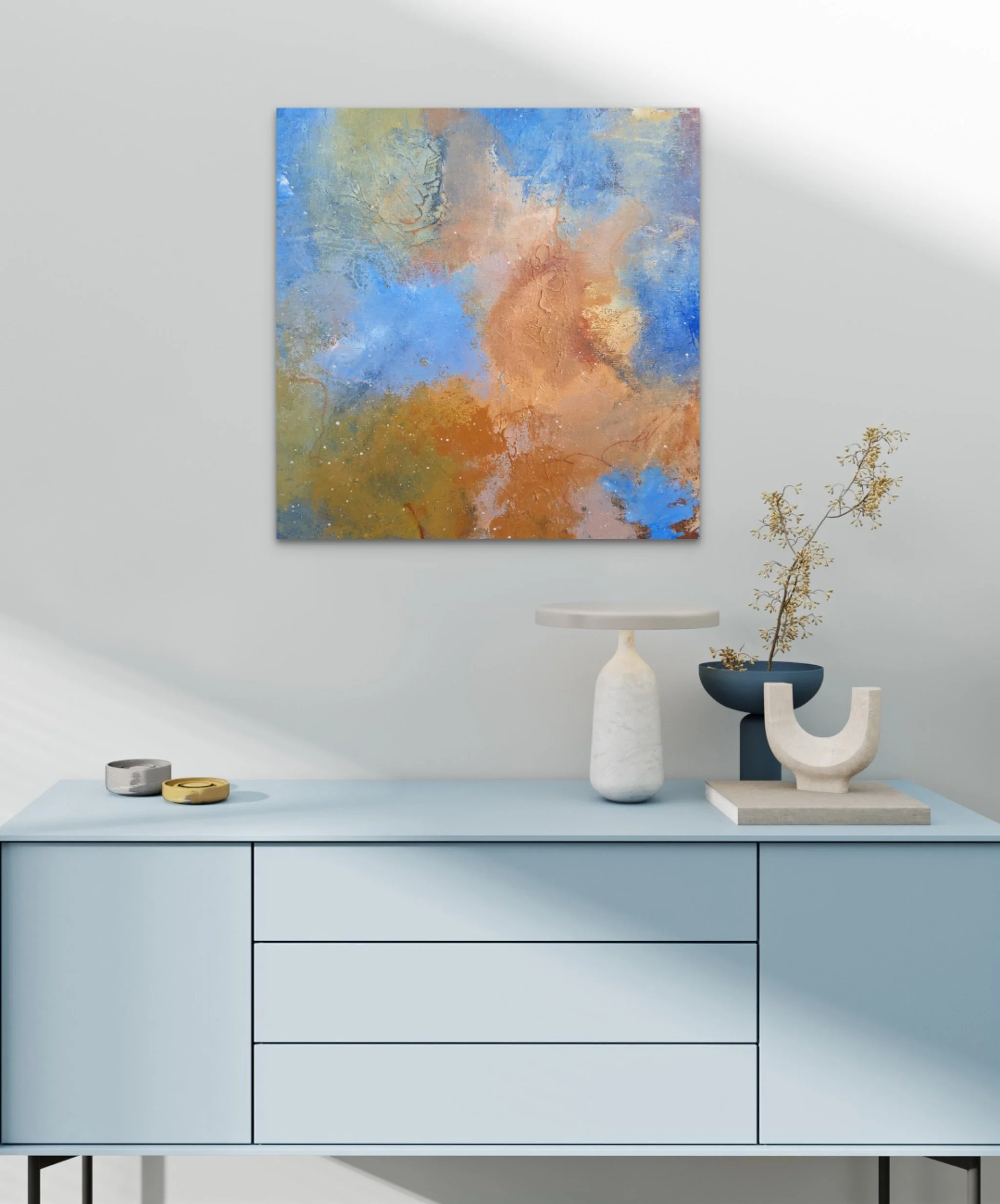 Modern blue cabinet with a light blue surface, decorated with a tall white marble vase with a branch of gold leaves, a abstract sculpture with a black ball and white base, and a white ceramic dish with a black container. Abstract painting with blue, beige, and orange tones hangs on a white wall behind.