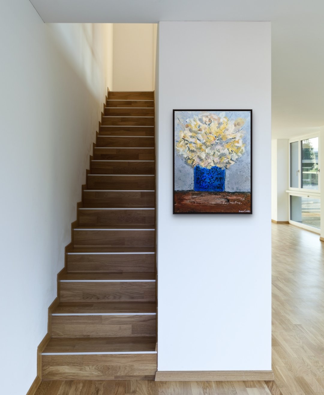 Interior view of a modern home featuring a wooden staircase and a wall with an abstract floral painting in a black frame, with windows letting in natural light.