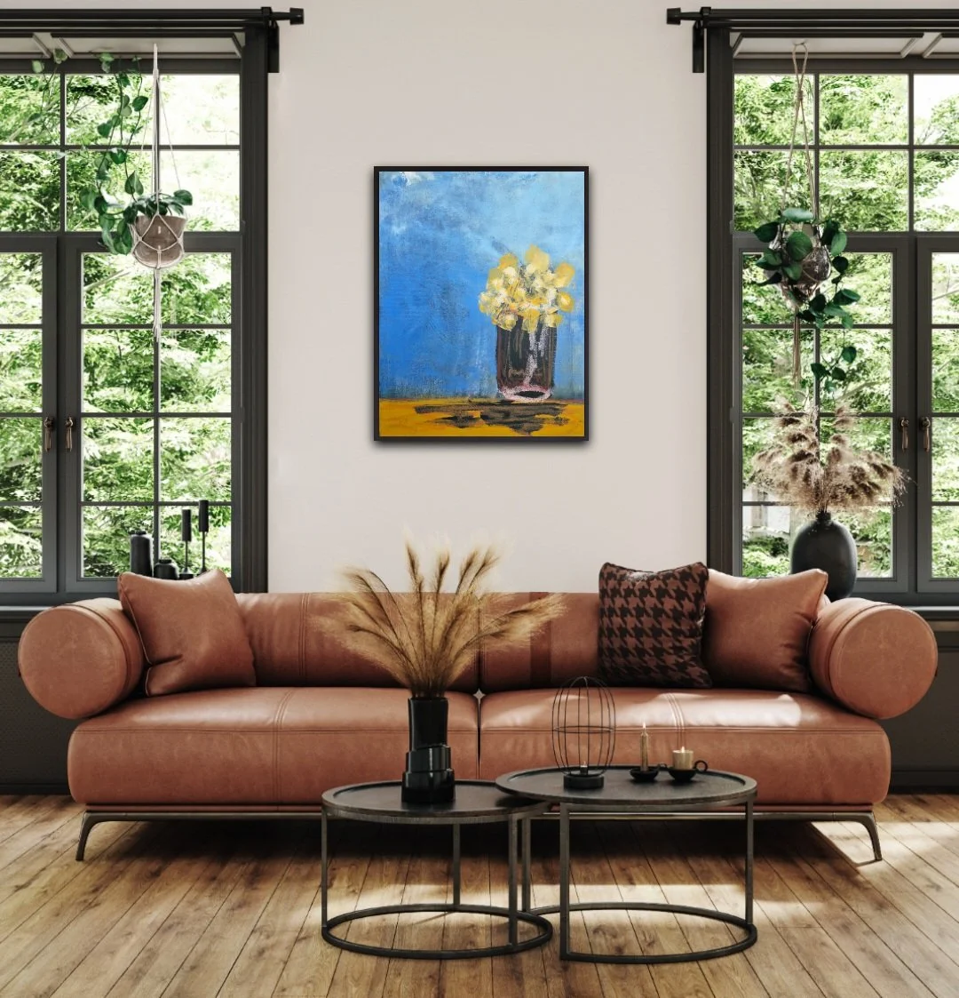 A cozy living room with a pink leather sofa, decorative pillows, and two black round coffee tables with decor items. Two large windows with black frames and hanging plants are behind the sofa. A colorful painting of a vase with flowers hangs on the white wall between the windows. The floor is wood, and the room is filled with natural light.