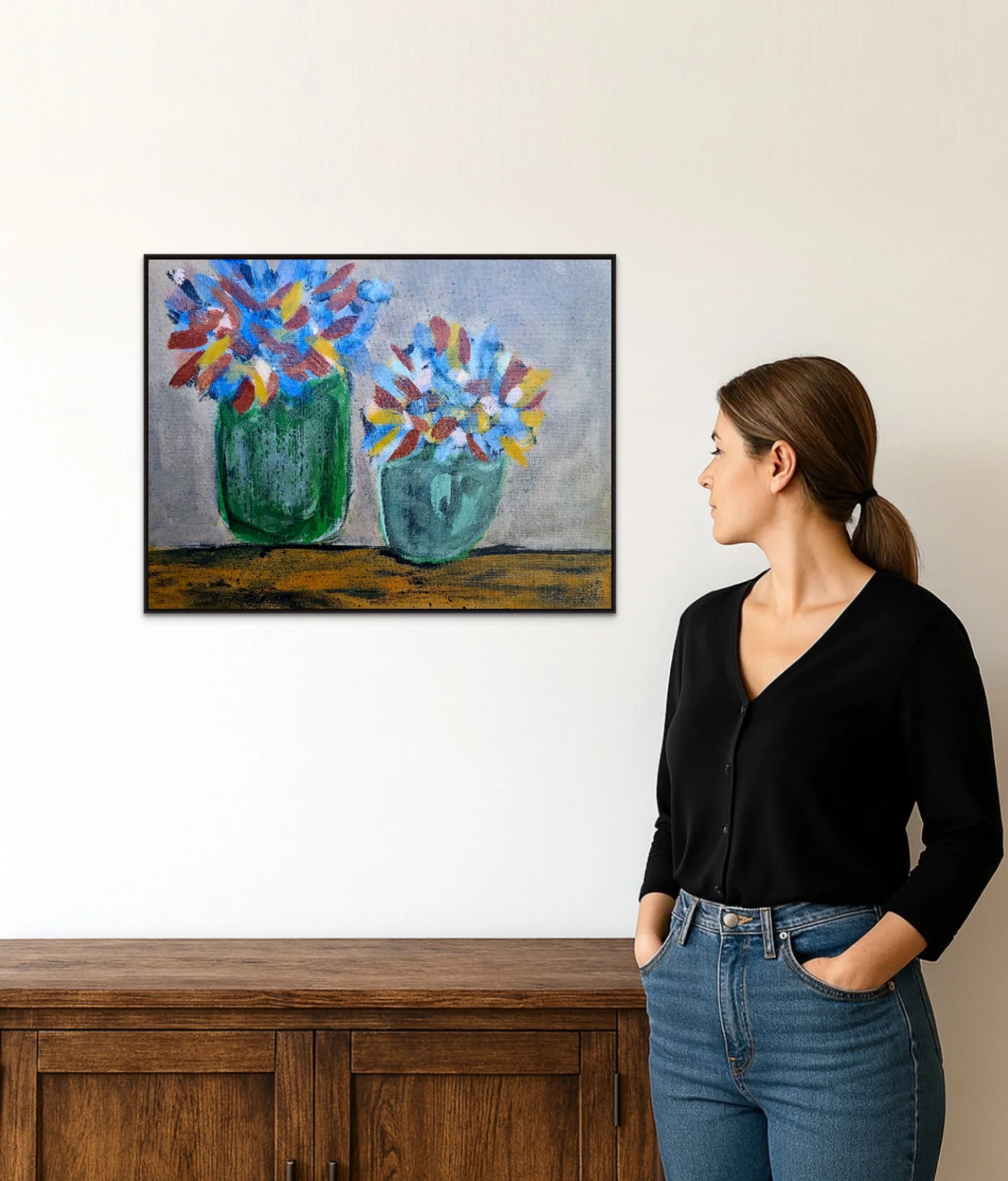 A woman with brown hair in a ponytail wearing a black blazer and blue jeans, standing indoors next to a wooden cabinet, with a traditional painting of colorful vases of flowers hanging on a white wall behind her.