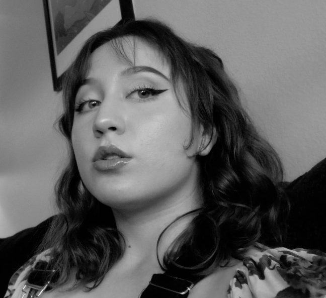 Black and white photo of a woman with shoulder-length wavy hair, wearing makeup with winged eyeliner and glossy lips, sitting on a couch, with a framed picture on the wall in the background.