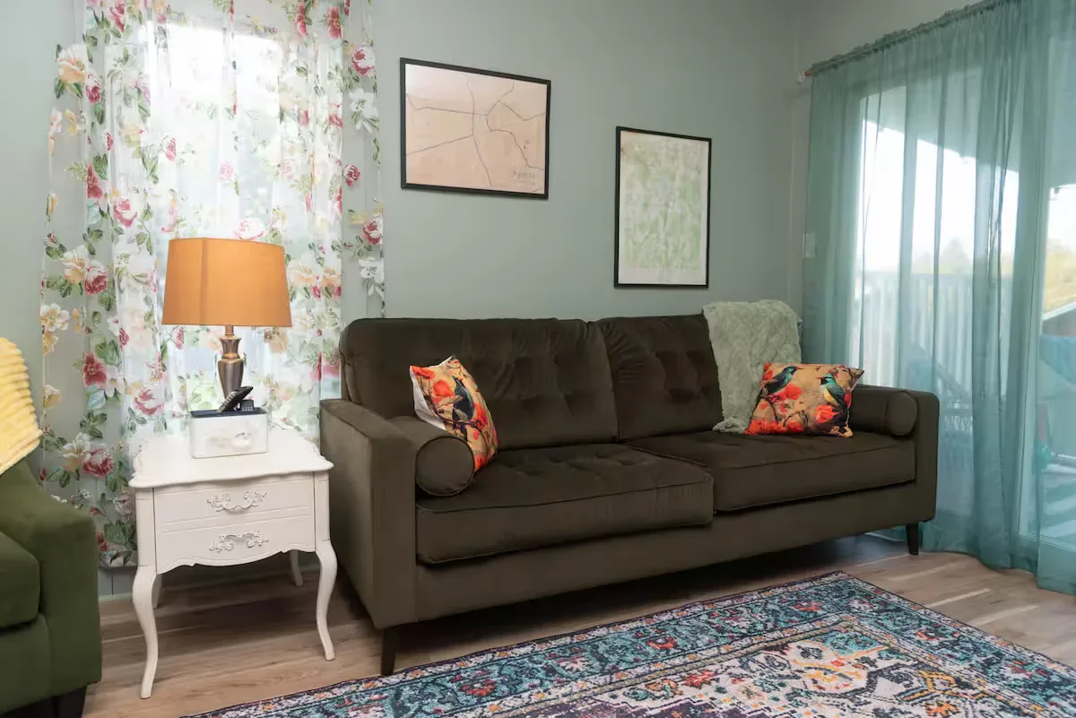 A living room with a dark green sofa, floral throw pillows, a white side table with a lamp, and framed wall art. The room has floral curtains and teal sheer curtains over a sliding glass door. An area rug is on the wooden floor.