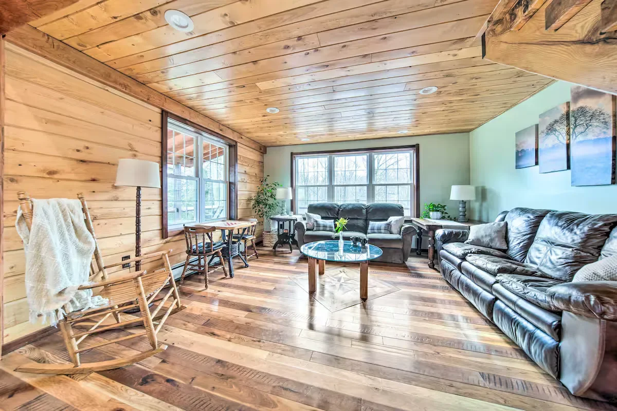 Living room with wooden walls and ceiling, hardwood floor, two leather sofas, a coffee table, a rocking chair with a blanket, side tables with lamps, and potted plants near windows.