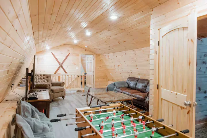 Attic game room with foosball table, two couches, and a TV. Wooden paneling walls and ceiling, with a small railing and staircase in the background.