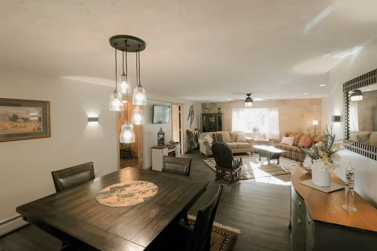 Living room with a dining area, featuring a dark wood dining table with chairs, a chandelier, a beige sofa, a rocking chair, a television, a cabinet, and a window with curtains.
